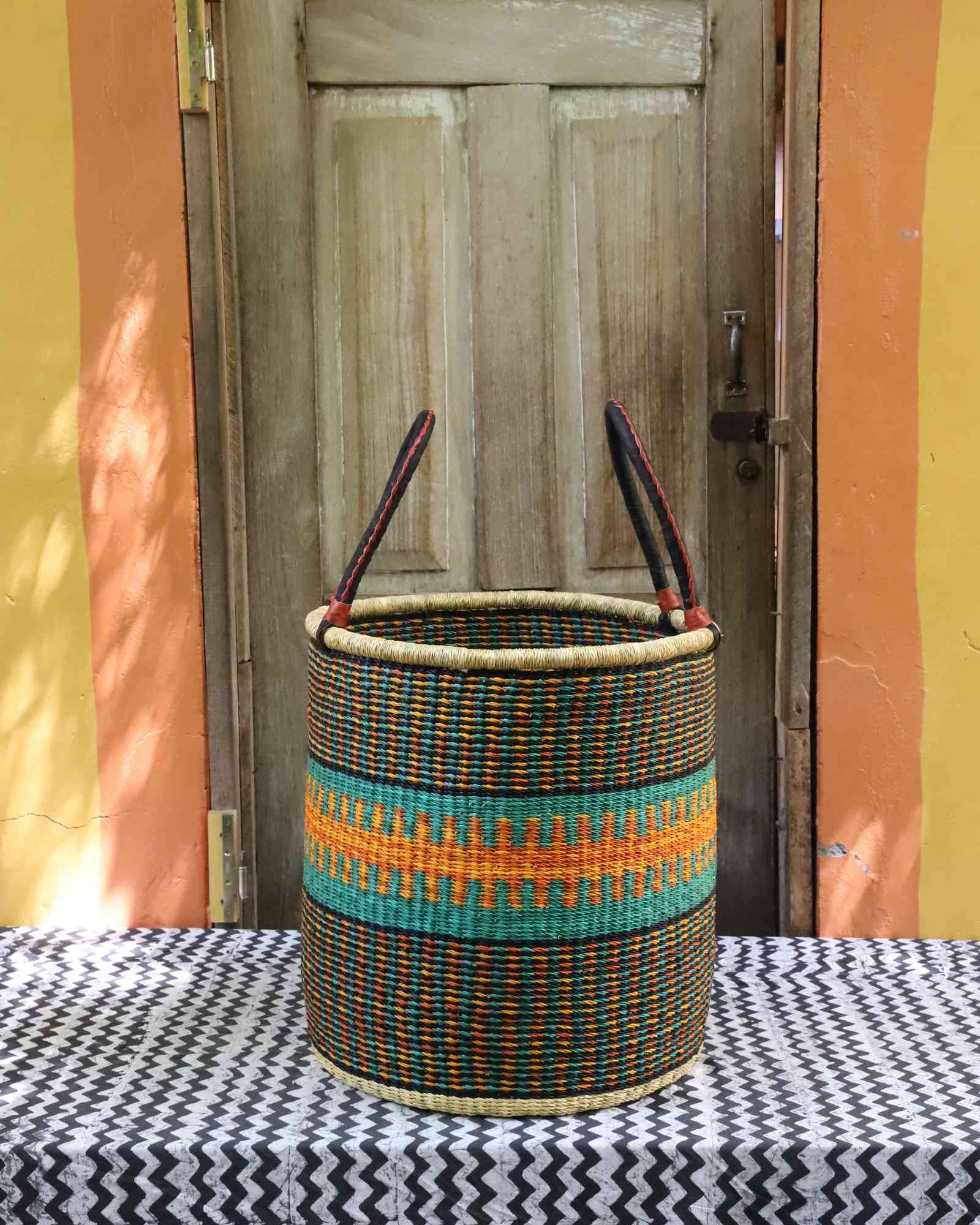 Tall open Bolga laundry basket in dark weave with a teal and orange patterned band and tall leather-topped handles.