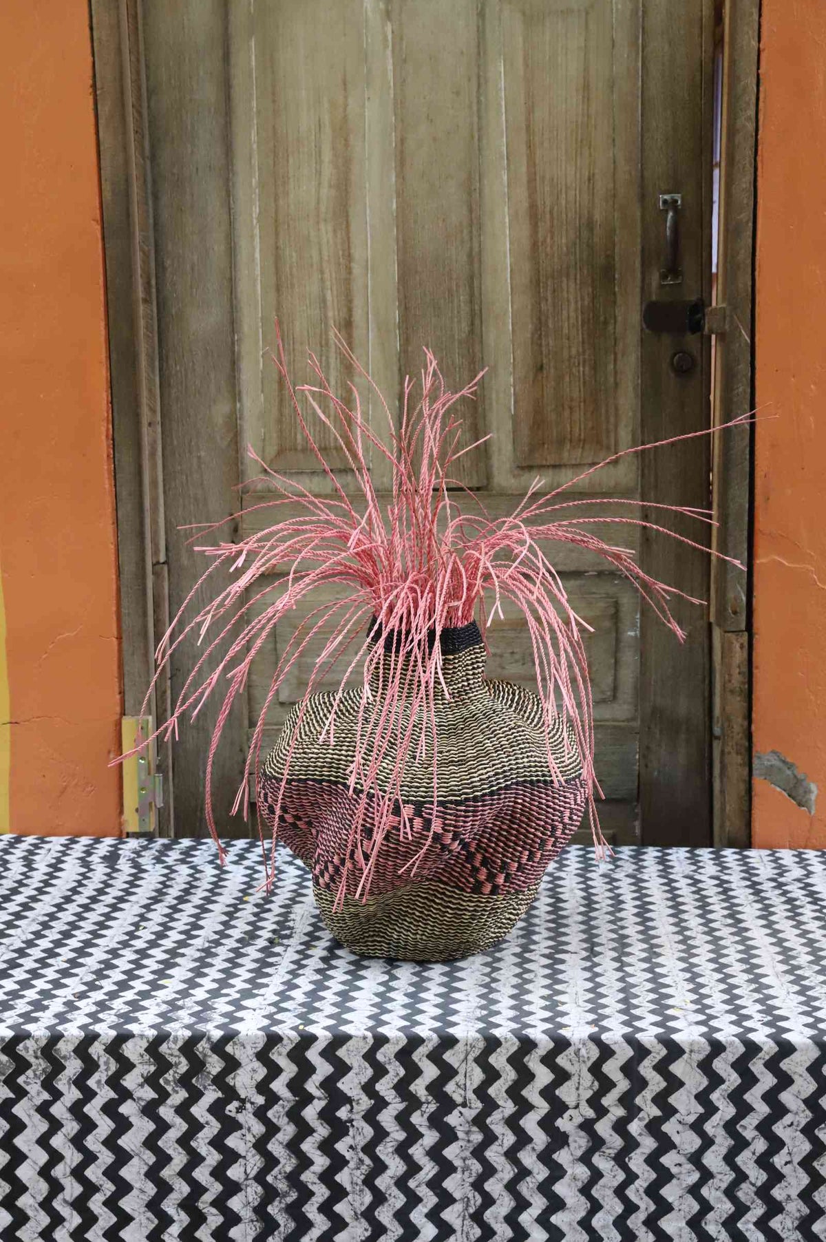 Handwoven Bolga Baobab-inspired basket in natural straw with a rust-red dotted band and pink grass sprigs.