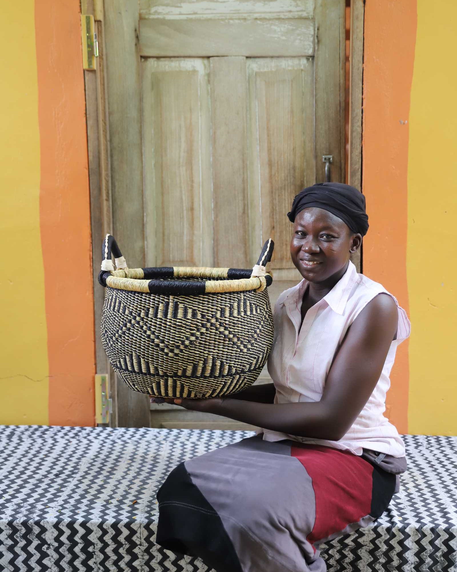 Handwoven Bolga storage basket in natural straw with black diamond pattern, held by a woman against a yellow wall.