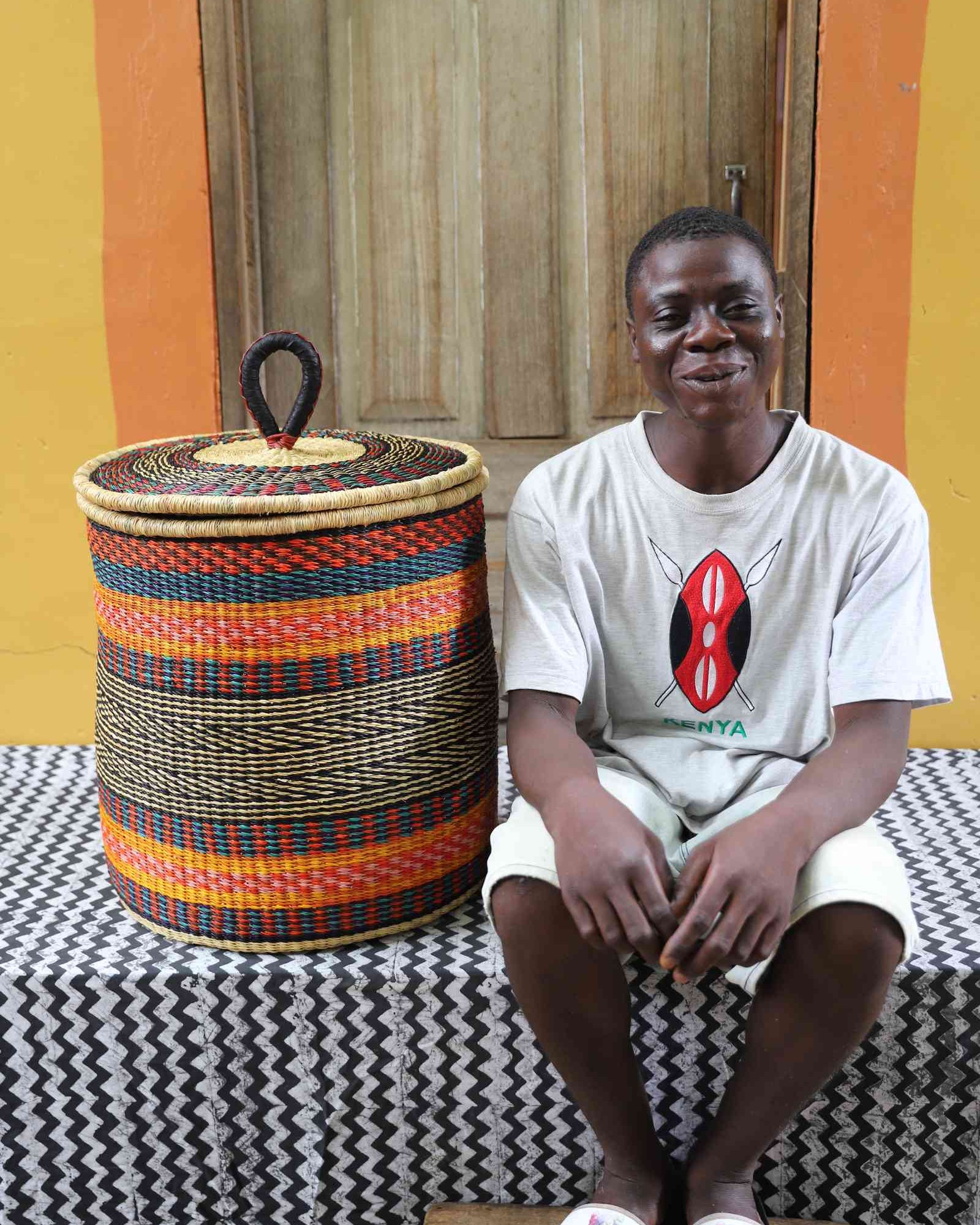 Coiled-rim lidded laundry basket in orange, red and green striped weave, shown beside a seated person for scale.