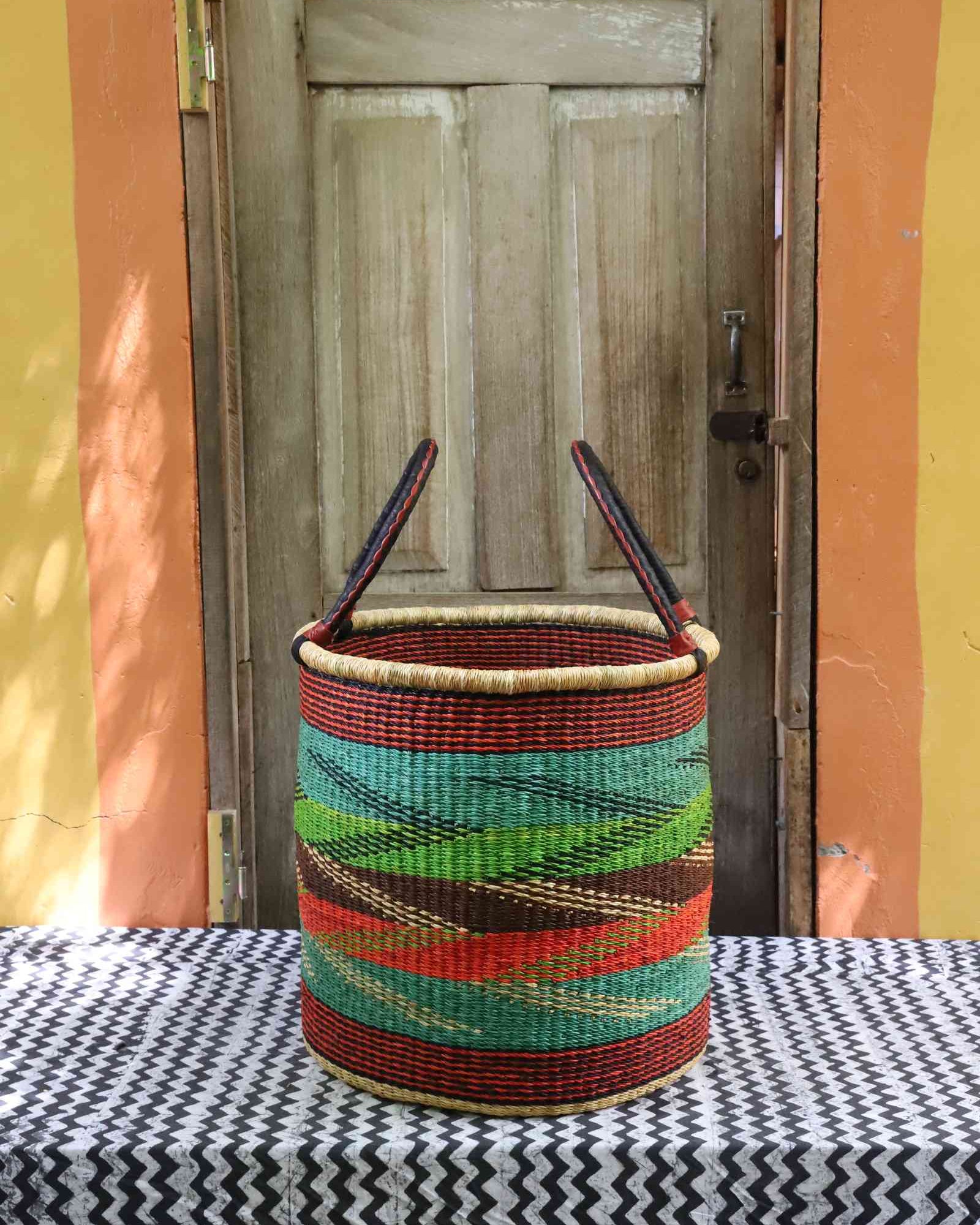 Tall open Bolga laundry basket with a multicolour diagonal striped weave in teal, green and rust, on a tablecloth.