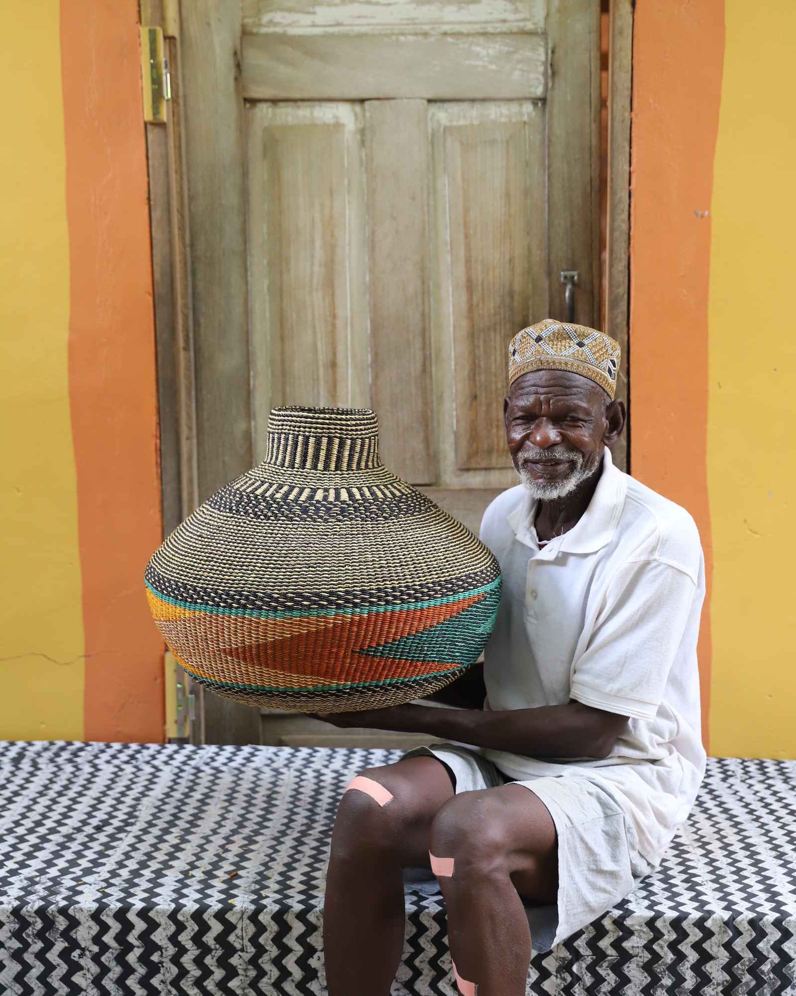 Handwoven African art basket in vase form with terracotta and teal bands, held by a man seated in Bolgatanga.