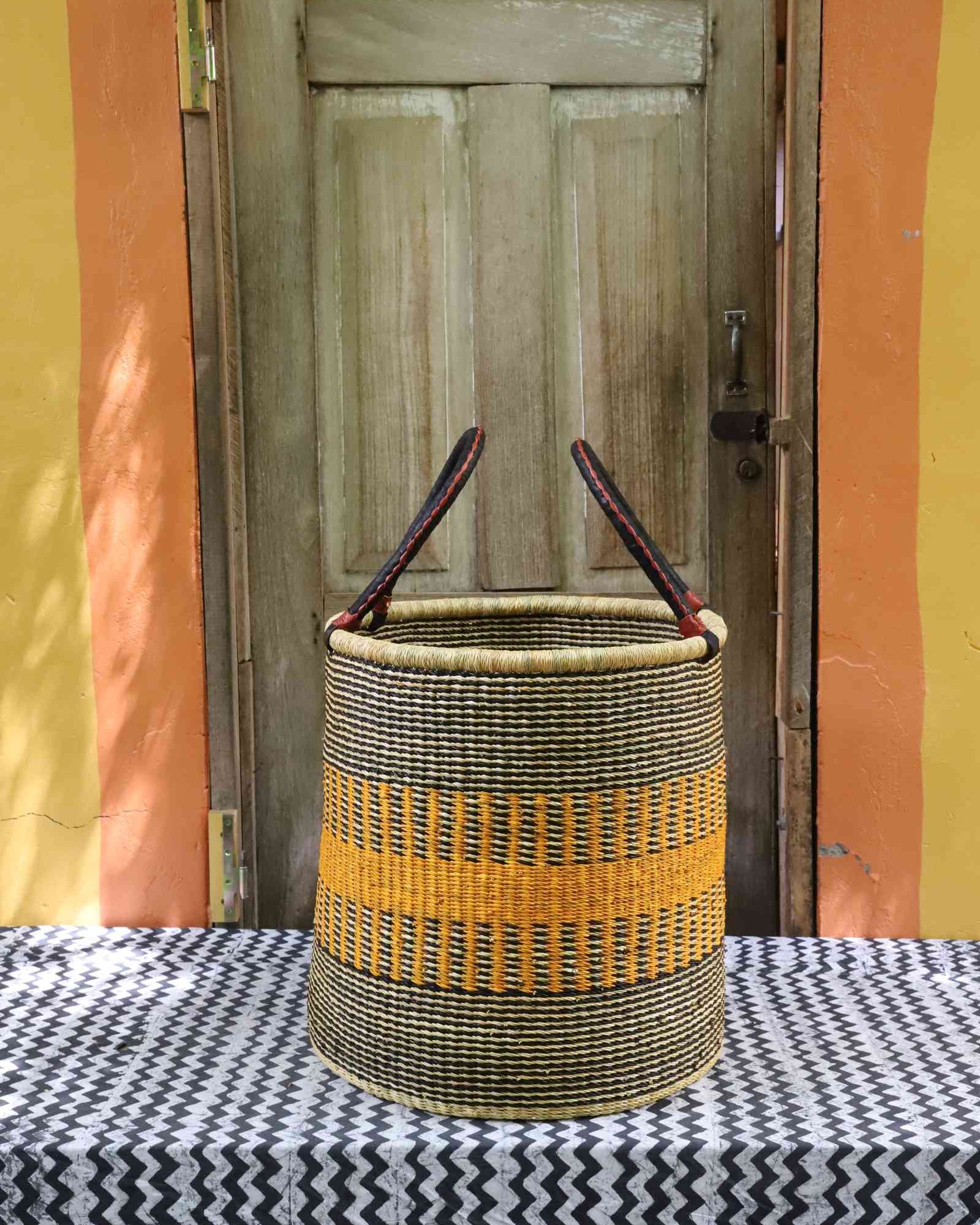 Tall open Bolga laundry basket in black and cream weave with a bold yellow stripe band and leather-topped handles.