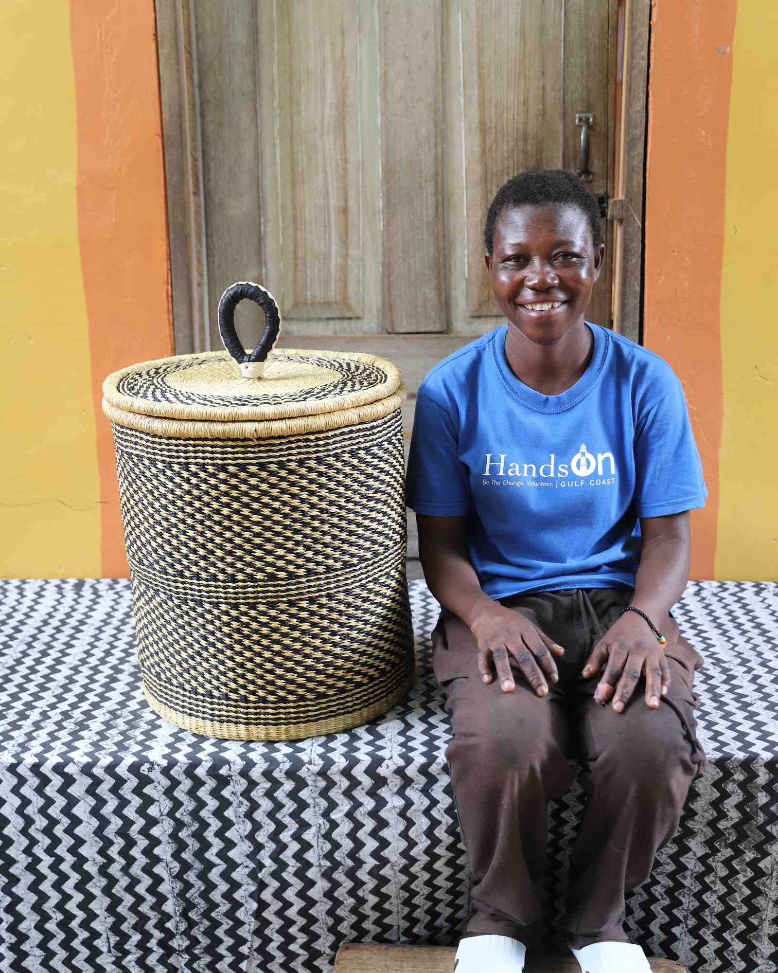 Coiled-rim lidded laundry basket in a black and tan speckled weave, shown beside a seated person for scale.