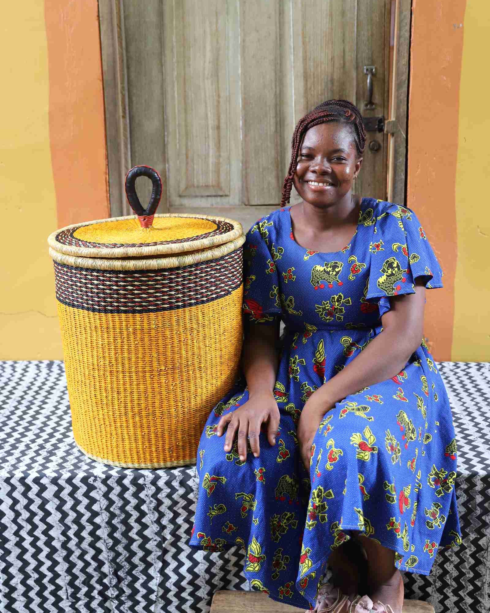 Coiled-rim lidded laundry basket in solid mustard weave, shown beside a seated person for scale.