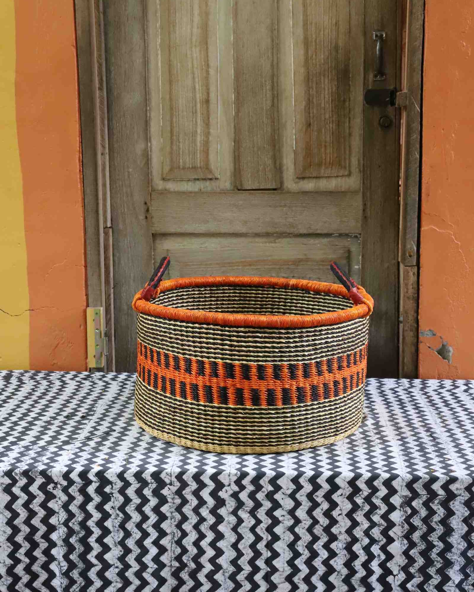 Handwoven Bolga storage basket in natural straw with an orange and black striped band and twin handles.