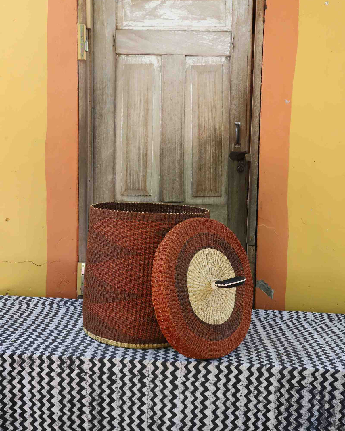 Handwoven Bolga laundry basket with lid removed, rust-red body with natural rim and matching lid.