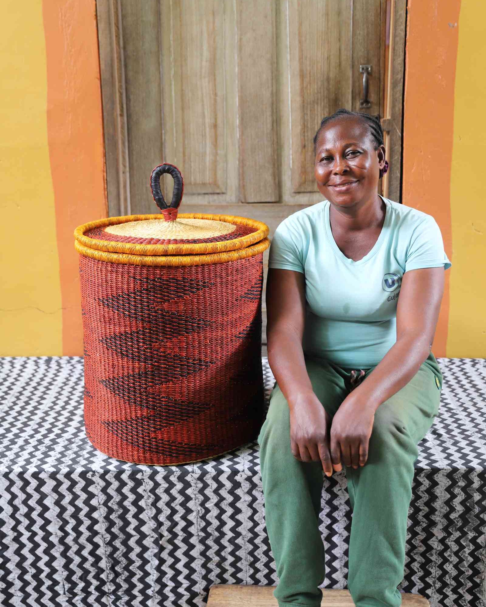 Coiled-rim lidded laundry basket in rust weave with a black diamond pattern, shown beside a seated person for scale.