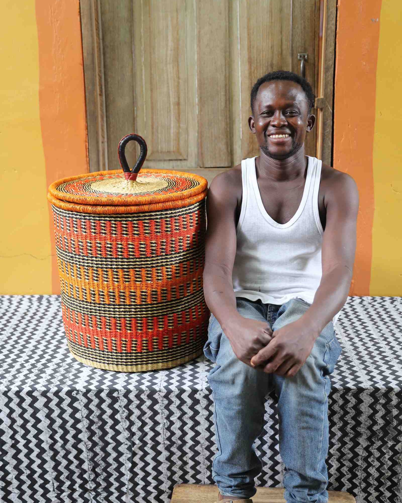 Coiled-rim lidded laundry basket in orange and black fine-striped weave, shown beside a seated person for scale.