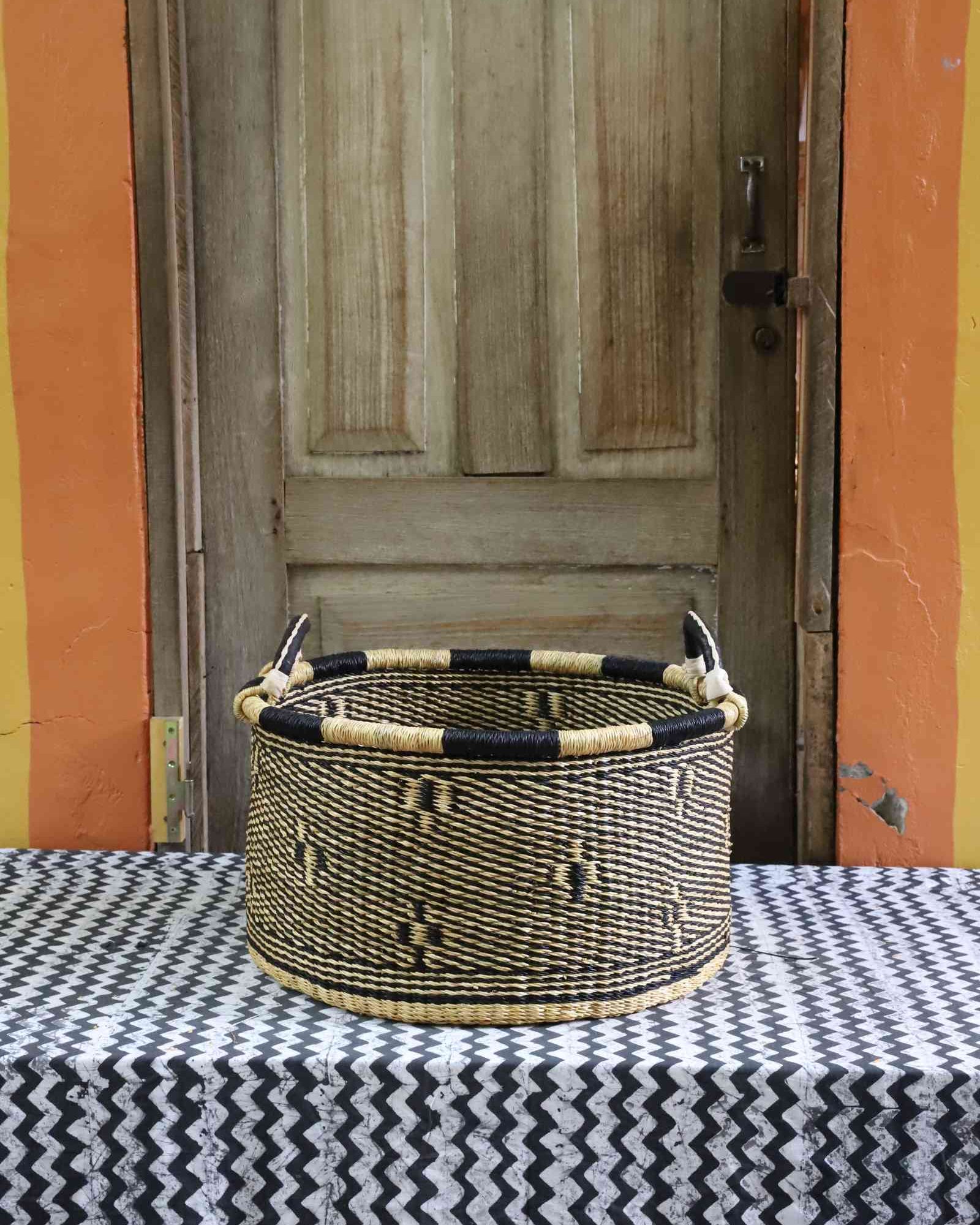 Handwoven Bolga storage basket in natural straw with a subtle dark motif and black-and-cream banded rim.
