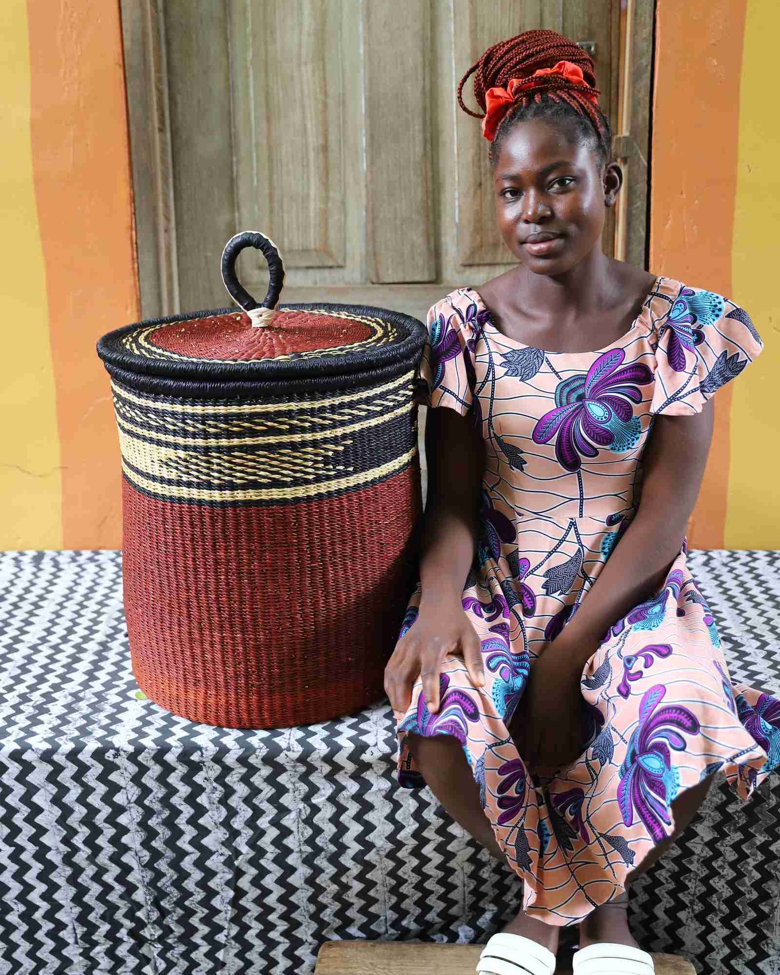 Coiled-rim lidded laundry basket in solid rust weave, shown beside a seated person for scale.