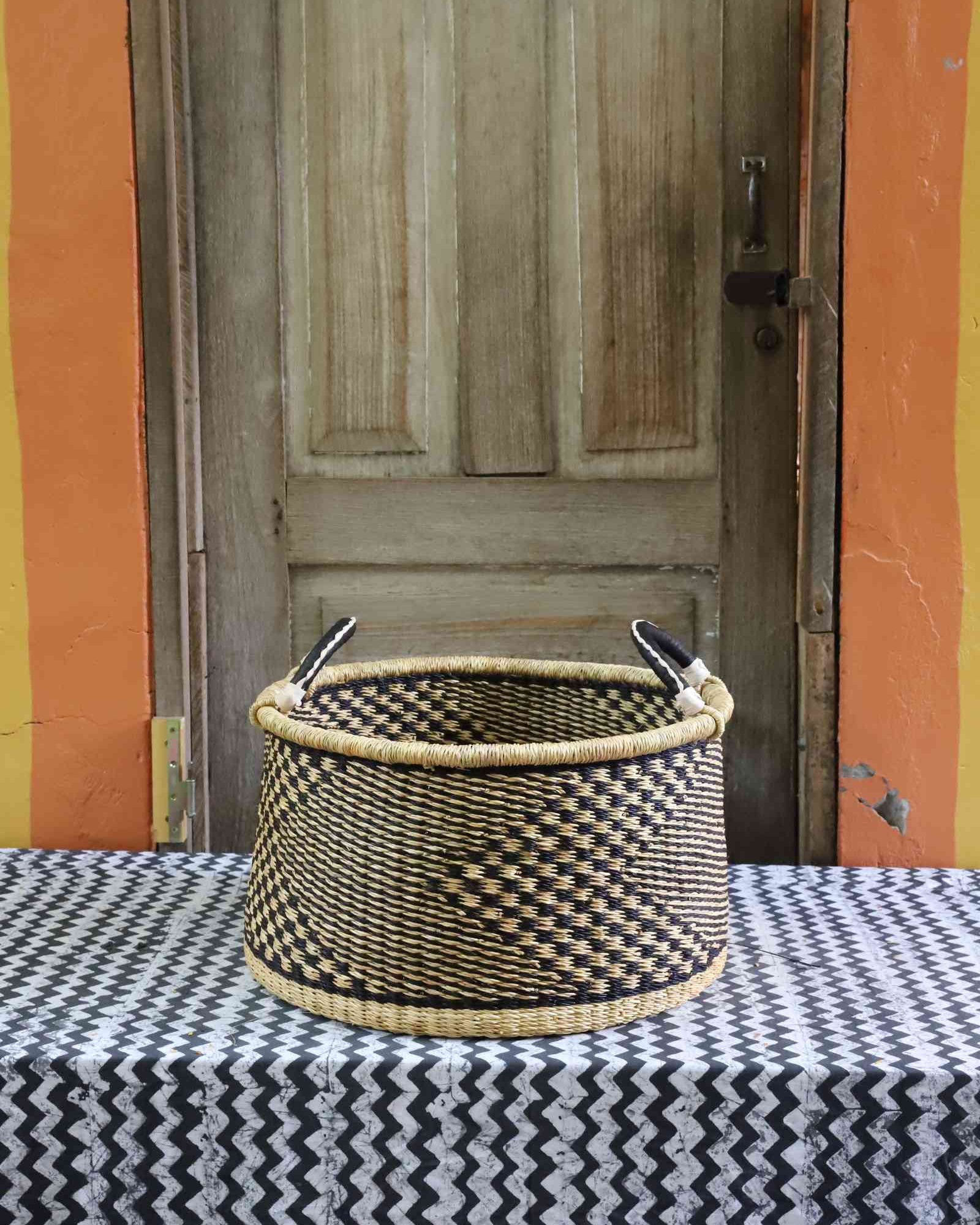 Handwoven Bolga storage basket in natural straw with a dark geometric weave and twin leather-wrapped handles.