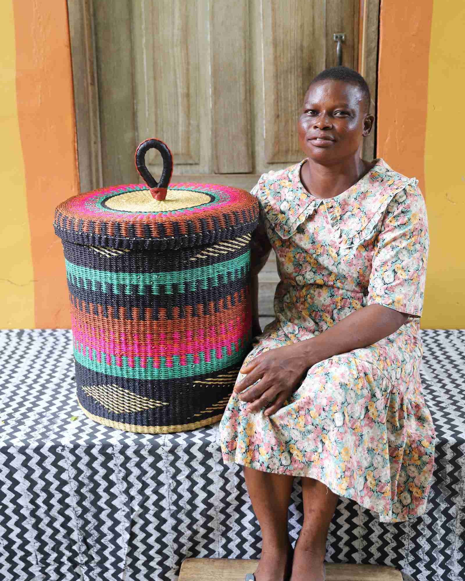 Lidded laundry basket in teal, pink and rust banded weave, shown beside a seated person for scale.