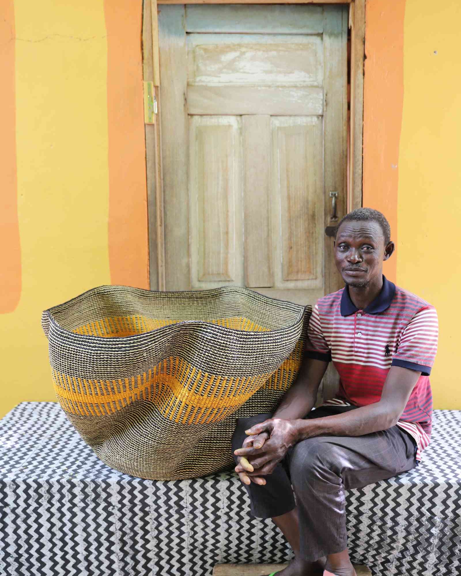 Large wave-form Bolga basket in grey and natural weave with an orange stripe, shown beside a seated man for scale.