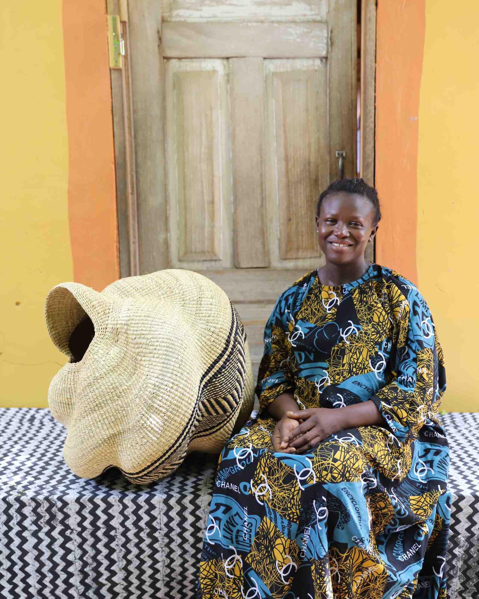 Natural straw Bolga basket in a spiral conch shape with a black geometric band, shown beside a seated woman for scale.