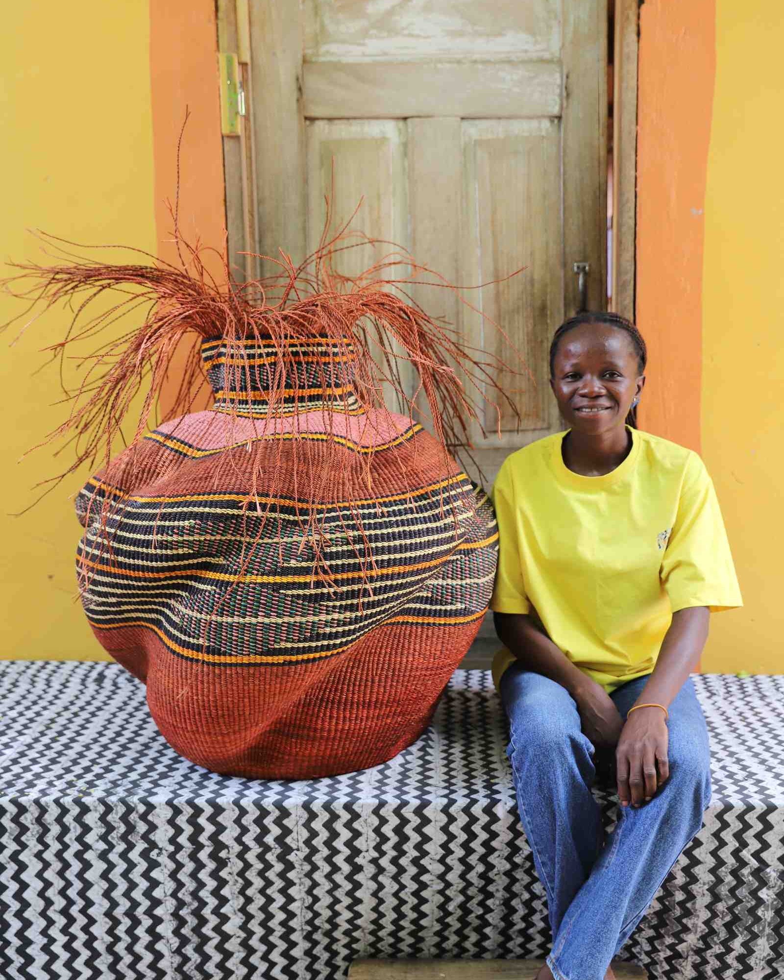 Large round Bolga basket in rust red with a striped band and loose straw fronds, shown beside a seated woman for scale.