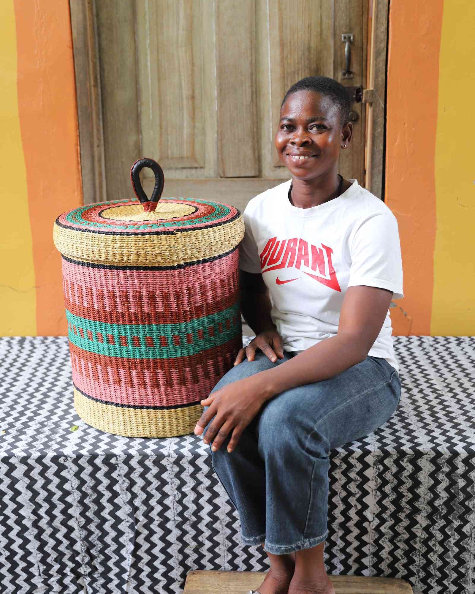 Lidded laundry basket in natural straw with pink and teal striped banding, shown beside a seated person for scale.