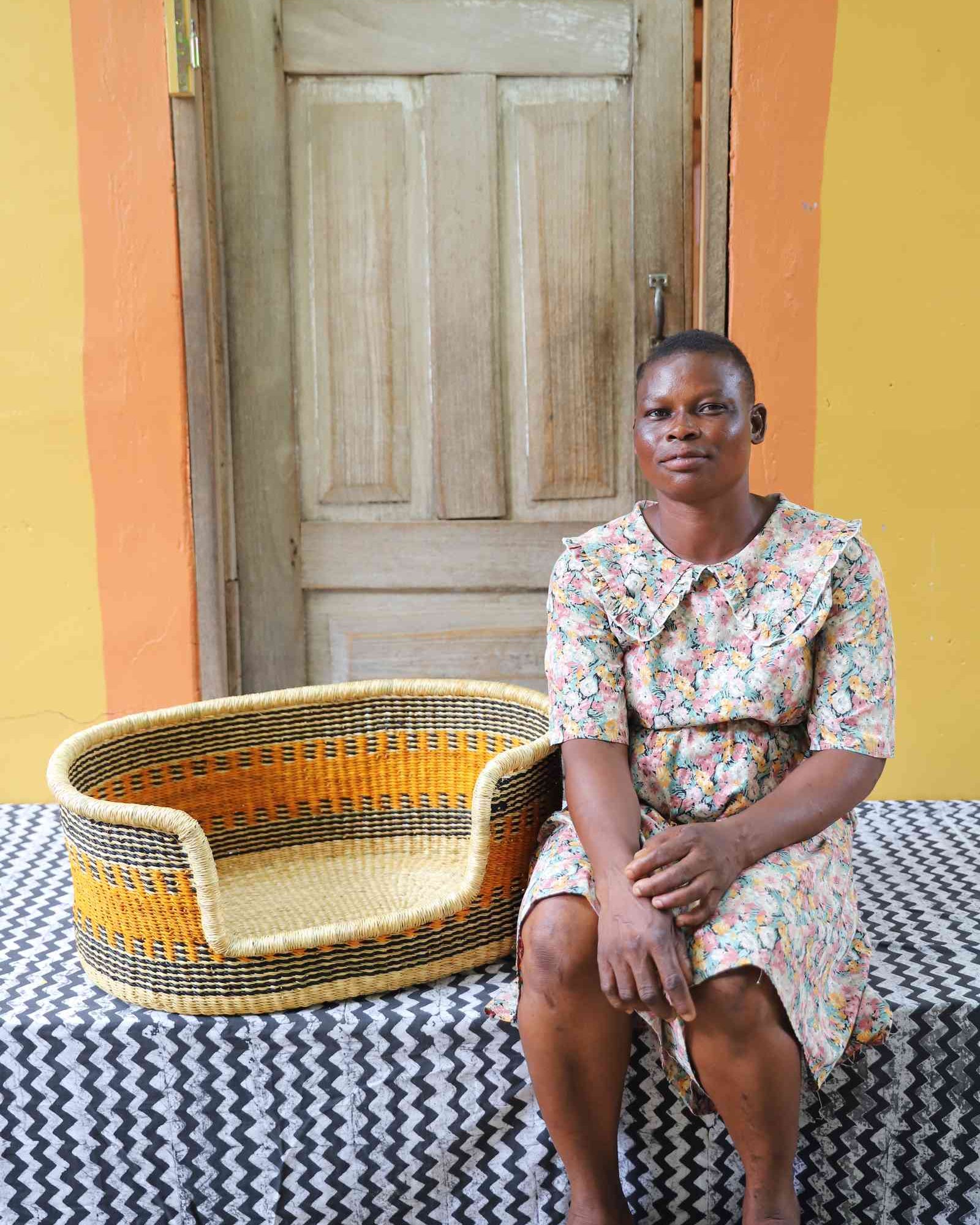 Woven dog bed in natural straw with an orange and dark checkered pattern, shown beside a seated person for scale.