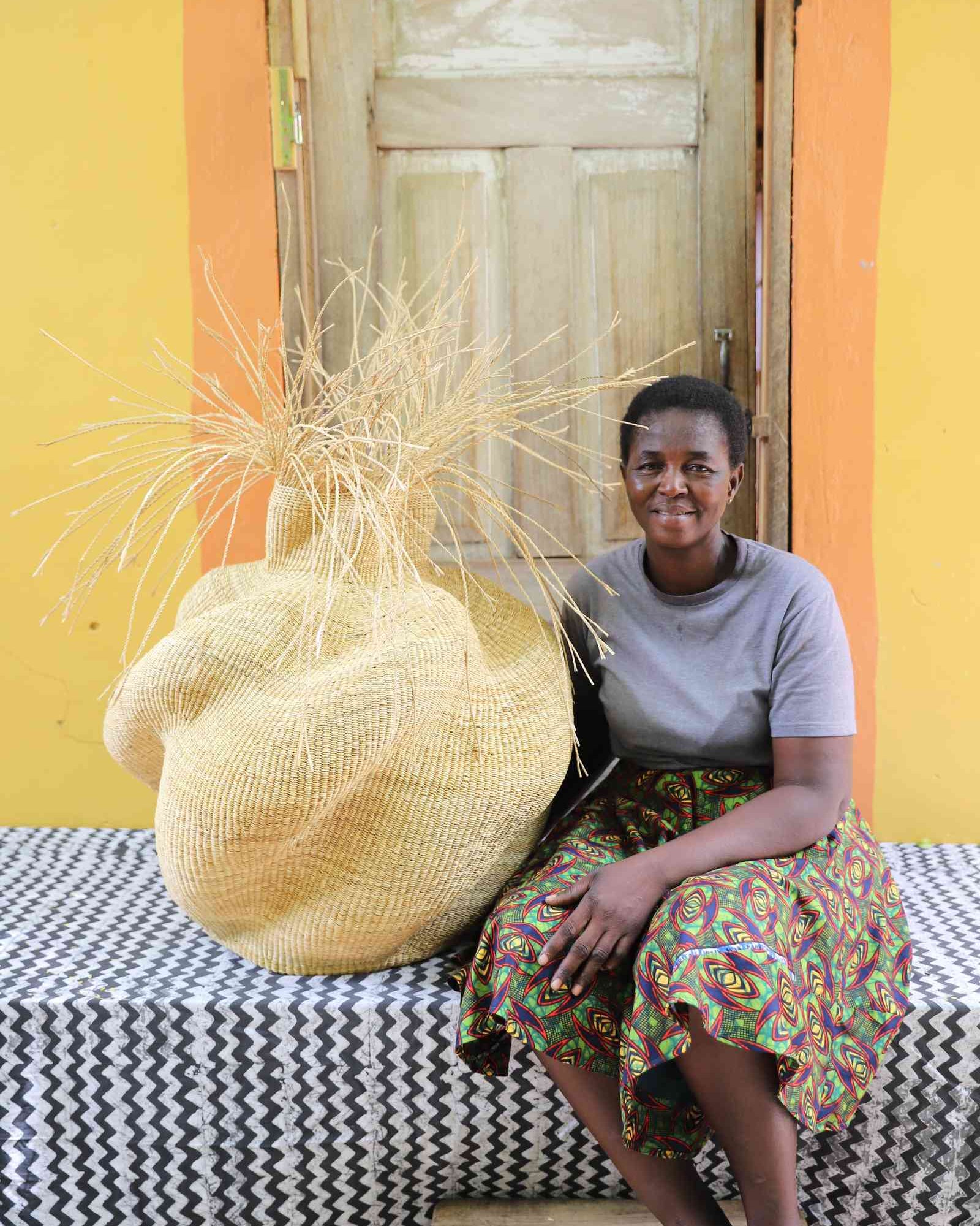 Round Bolga basket in natural straw with loose straw fronds at the neck, shown beside a seated woman for scale.