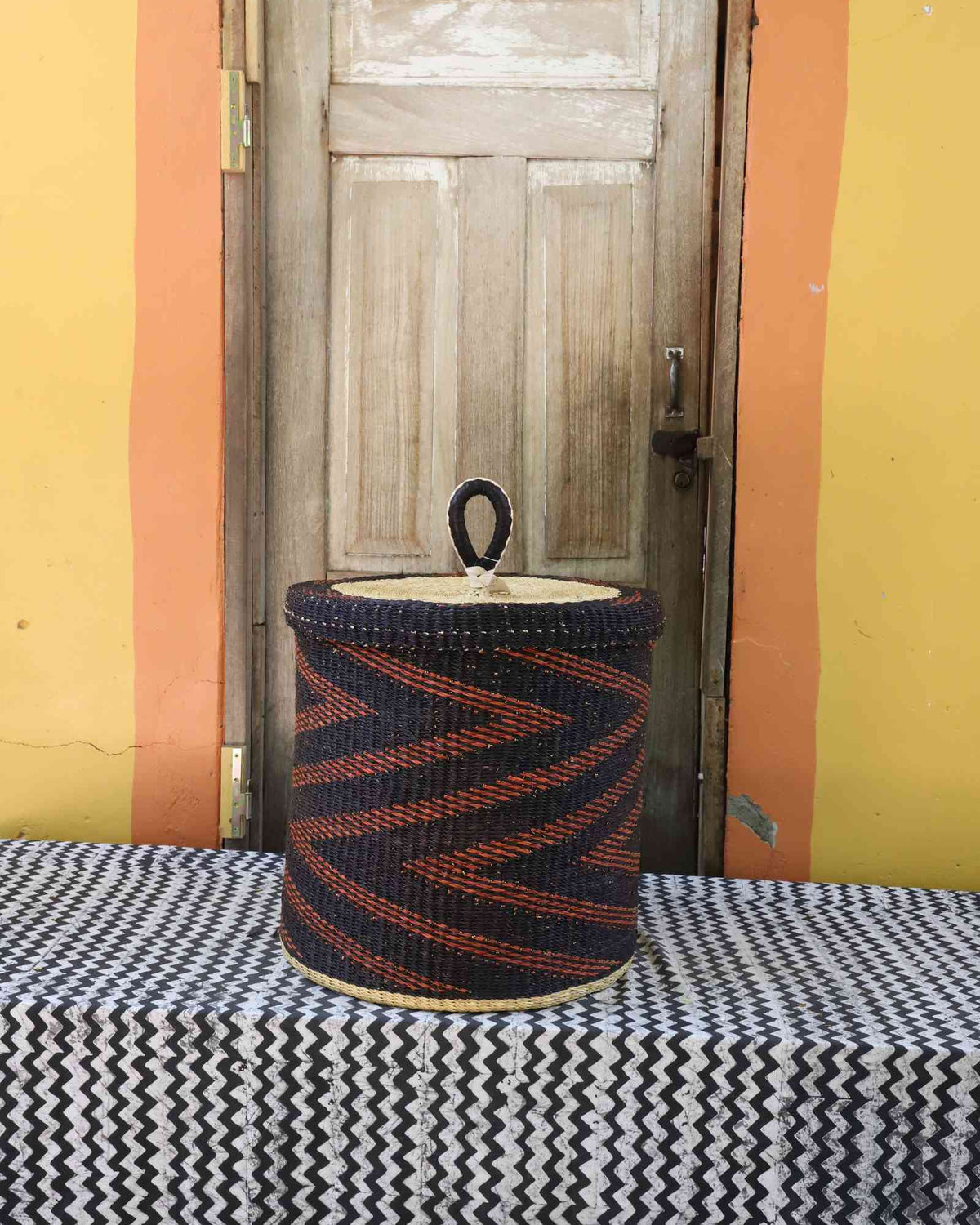 Handwoven Bolga laundry basket with lid in dark blue with a rust-orange zigzag arrow pattern.