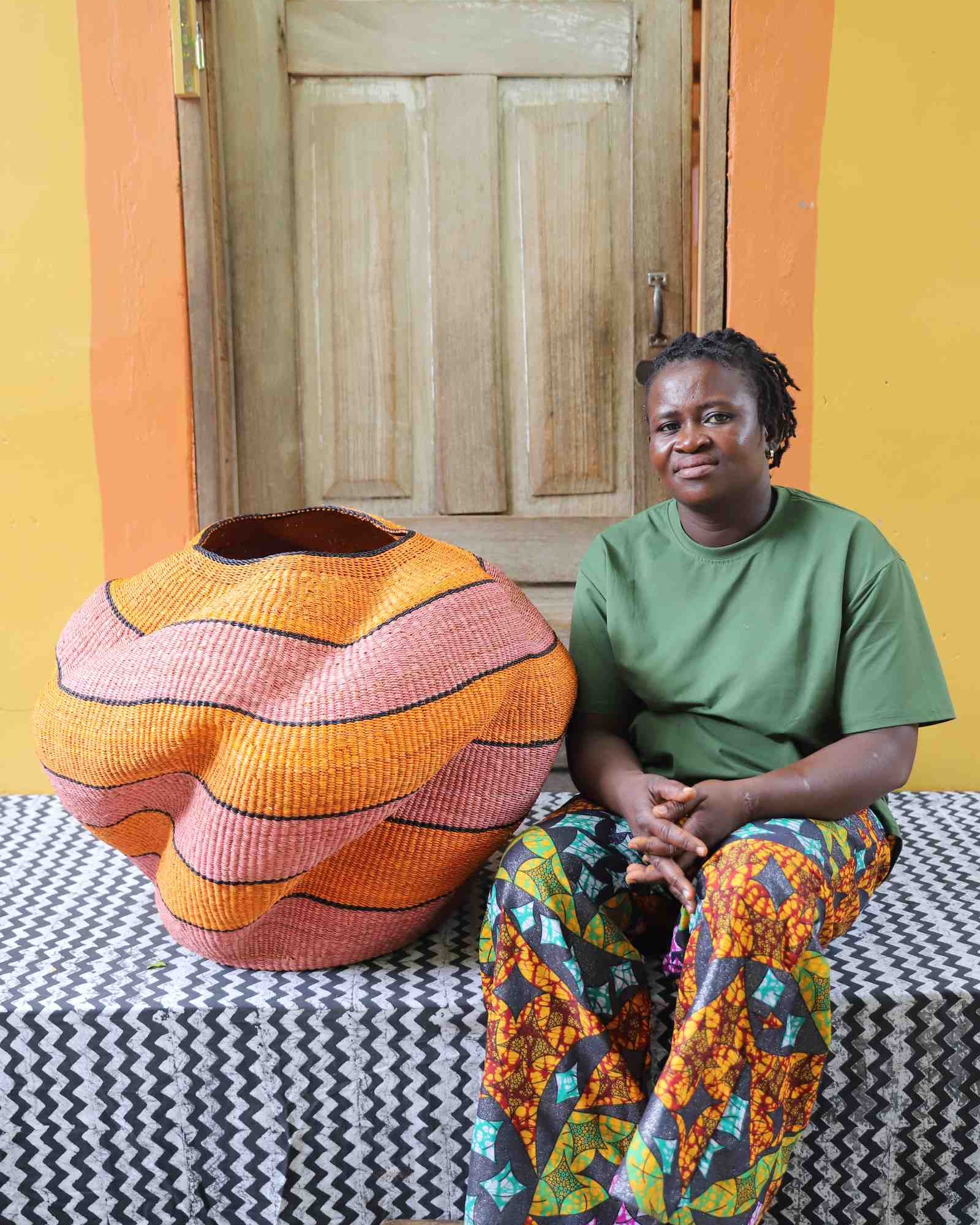 Large nest-shaped Bolga art basket in pink and rust wavy braided weave, shown beside a seated person for scale.