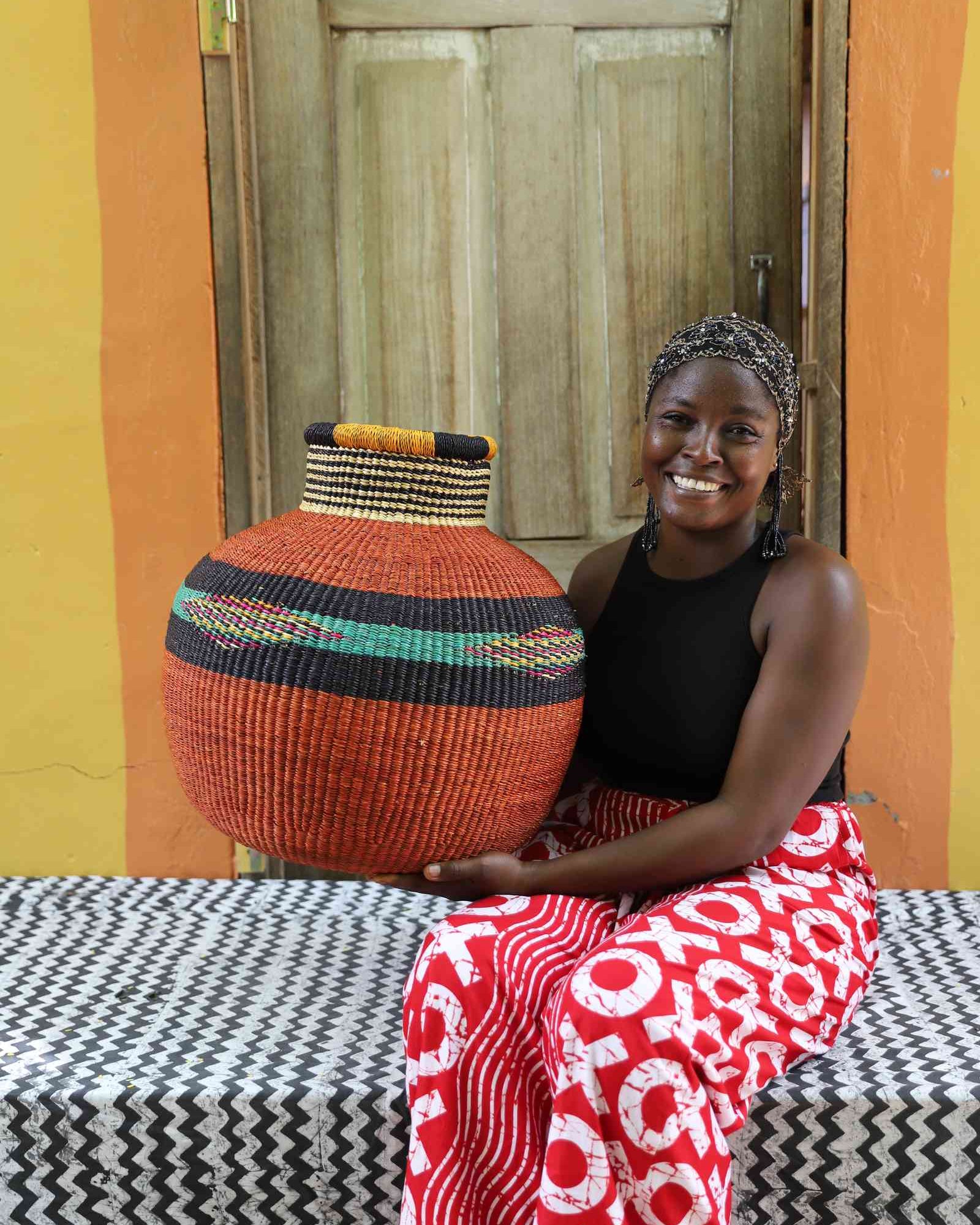Round terracotta Bolga basket with a striped neck and green diamond band, held by a seated woman for scale.