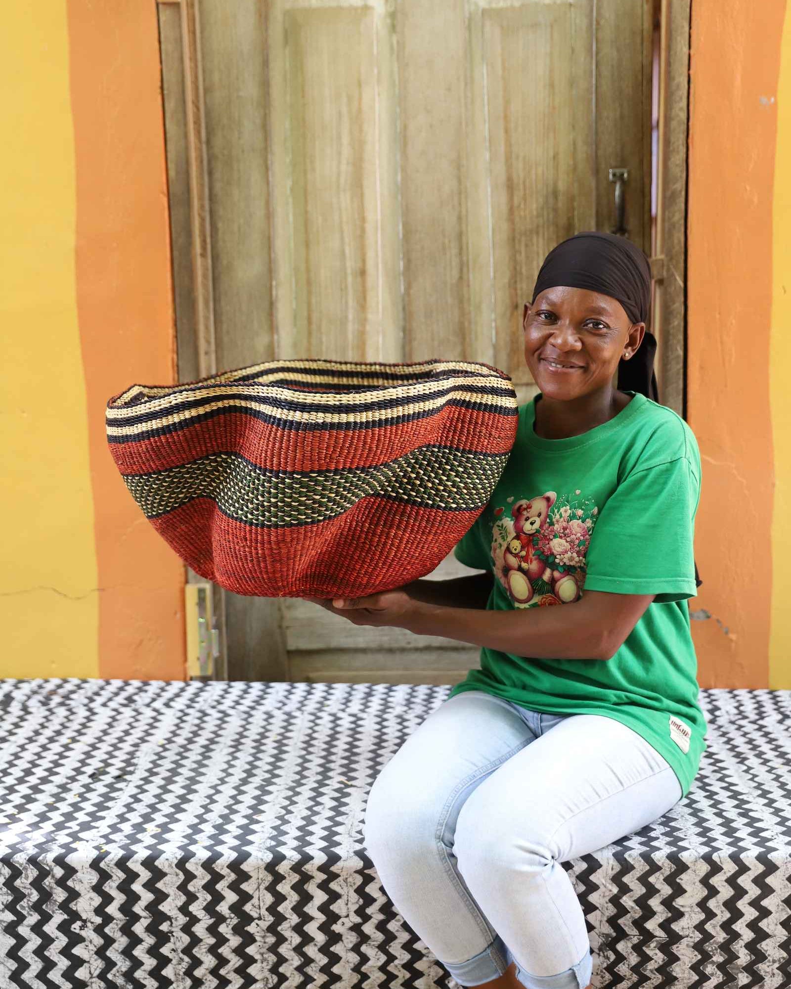 Wide rust red Bolga basket with a wavy rim and black-and-gold wave band, held by a seated woman for scale.