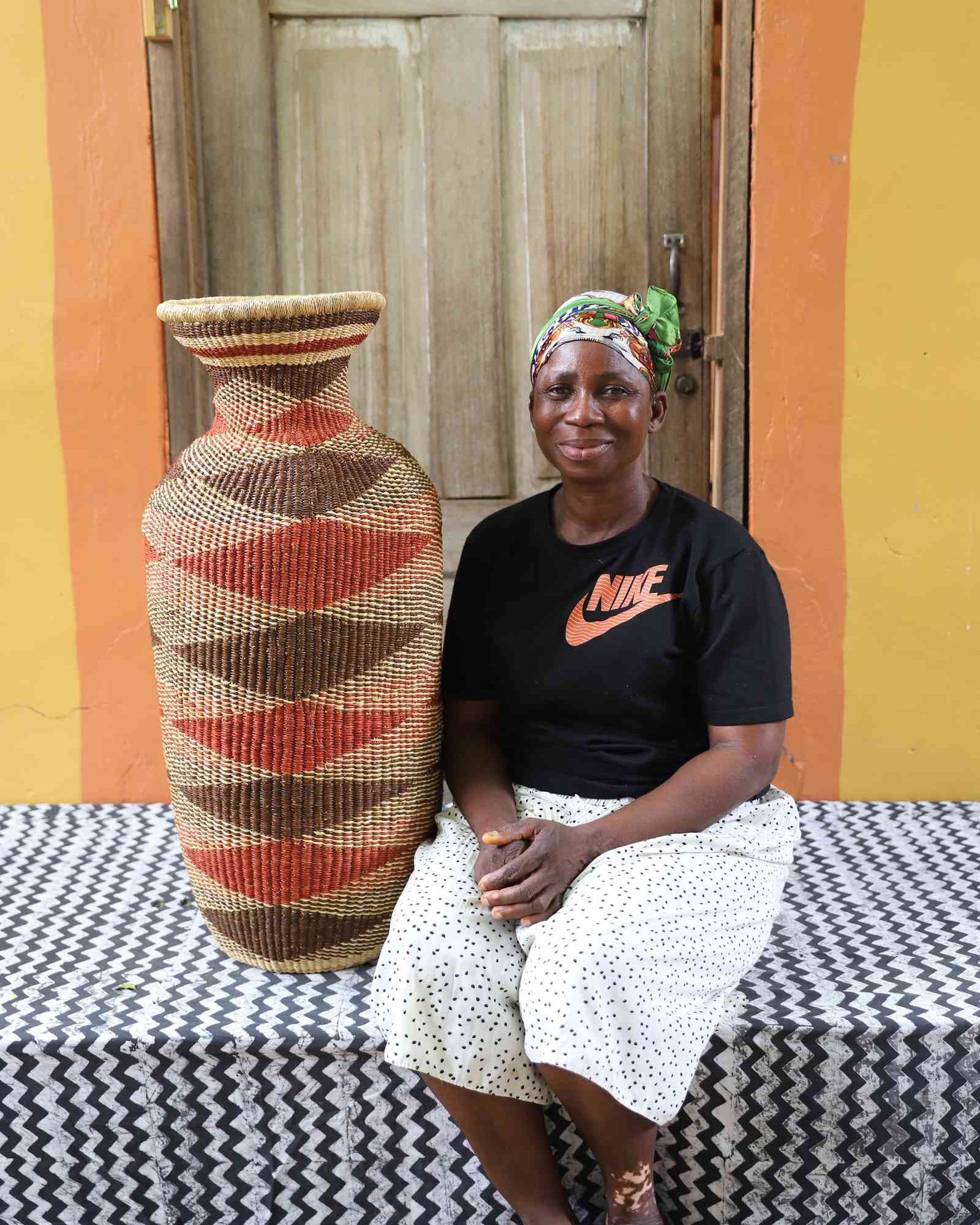 Tall vase-shaped Bolga art basket in natural straw with a brown and rust diamond pattern, shown beside a seated person for scale.