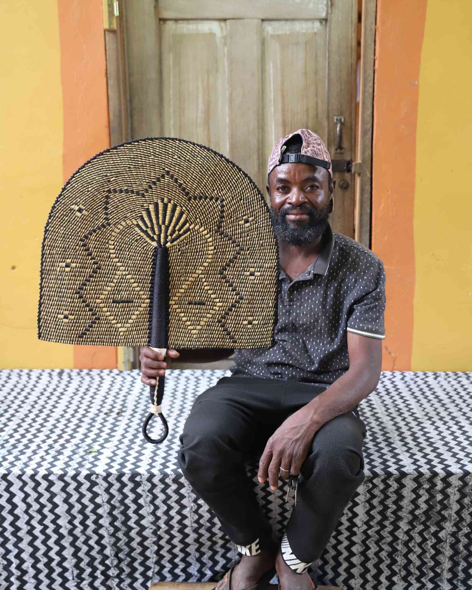 Naba Penga wall fan in dark brown straw with a tan star-shaped geometric weave, held by a seated person for scale.