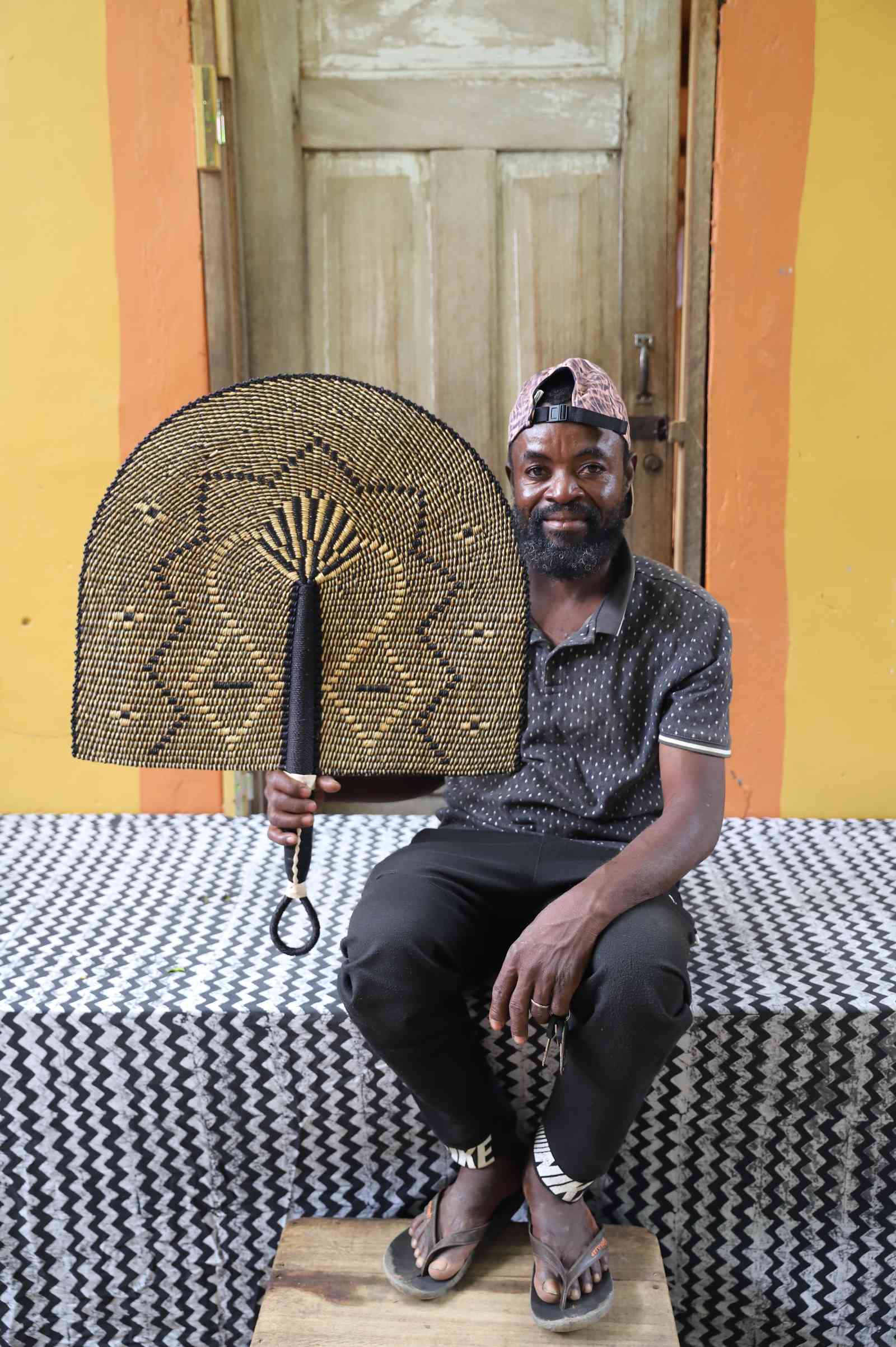 Naba Penga wall fan in dark brown straw with a tan star-shaped geometric weave, held by a seated person for scale.