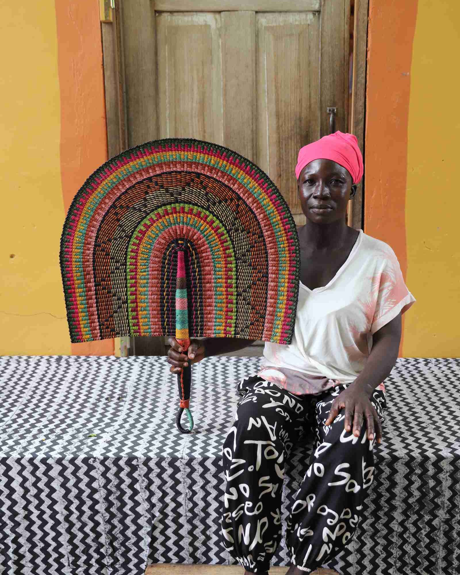 Naba Penga wall fan in natural straw with black, green and orange banding, held by a seated person for scale.