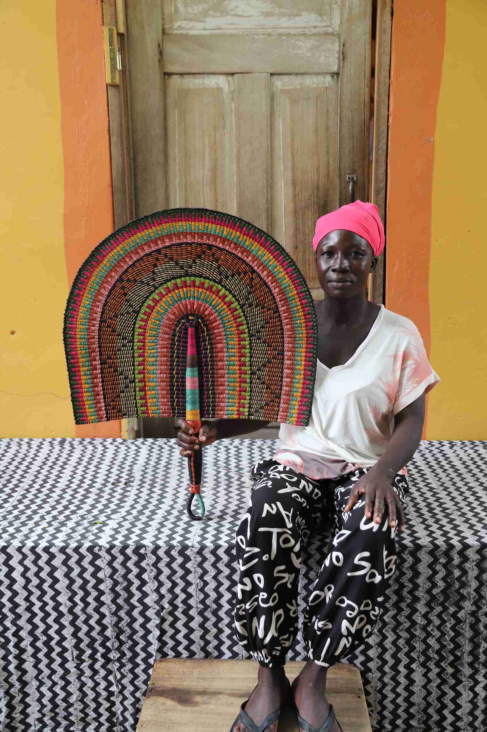 Naba Penga wall fan in natural straw with black, green and orange banding, held by a seated person for scale.