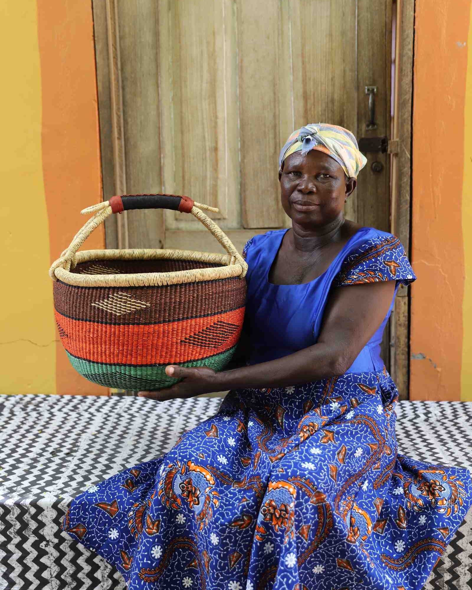 Round Bolga shopping basket striped in brown, orange and green with a leather-wrapped handle, held by a seated woman.