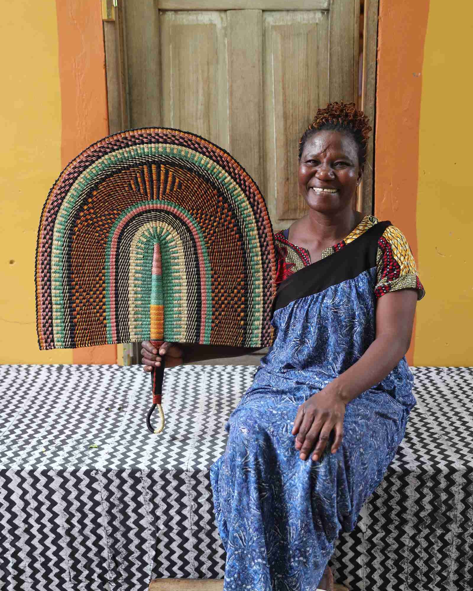 Naba Penga wall fan in natural straw with sage green and rust banded arches, held by a seated person for scale.