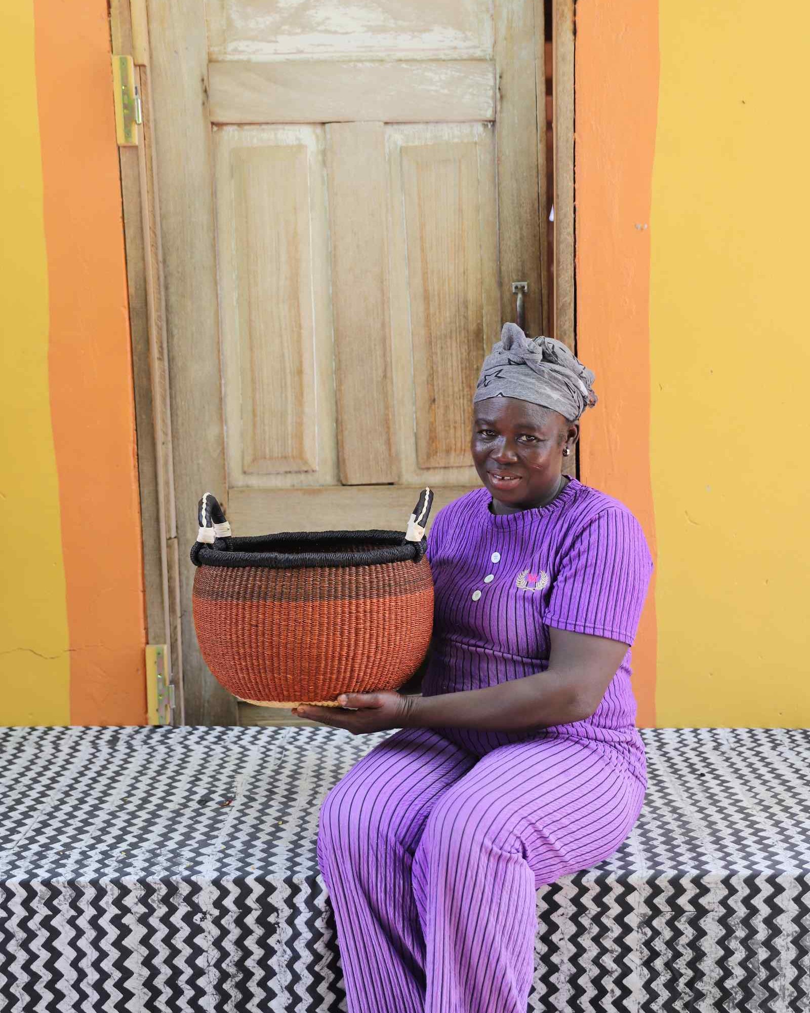 Handwoven Gbenka Bolga basket in rust with black rim, held by a woman seated against a yellow ochre wall in Bolgatanga.