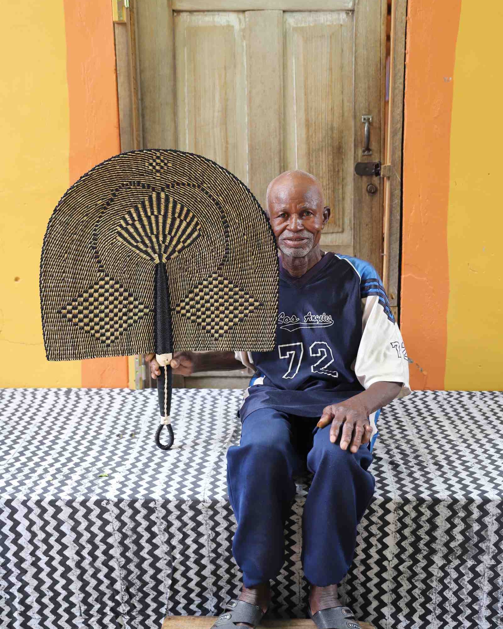 Naba Penga wall fan in natural straw with a dark geometric diamond weave, held by a seated person for scale.