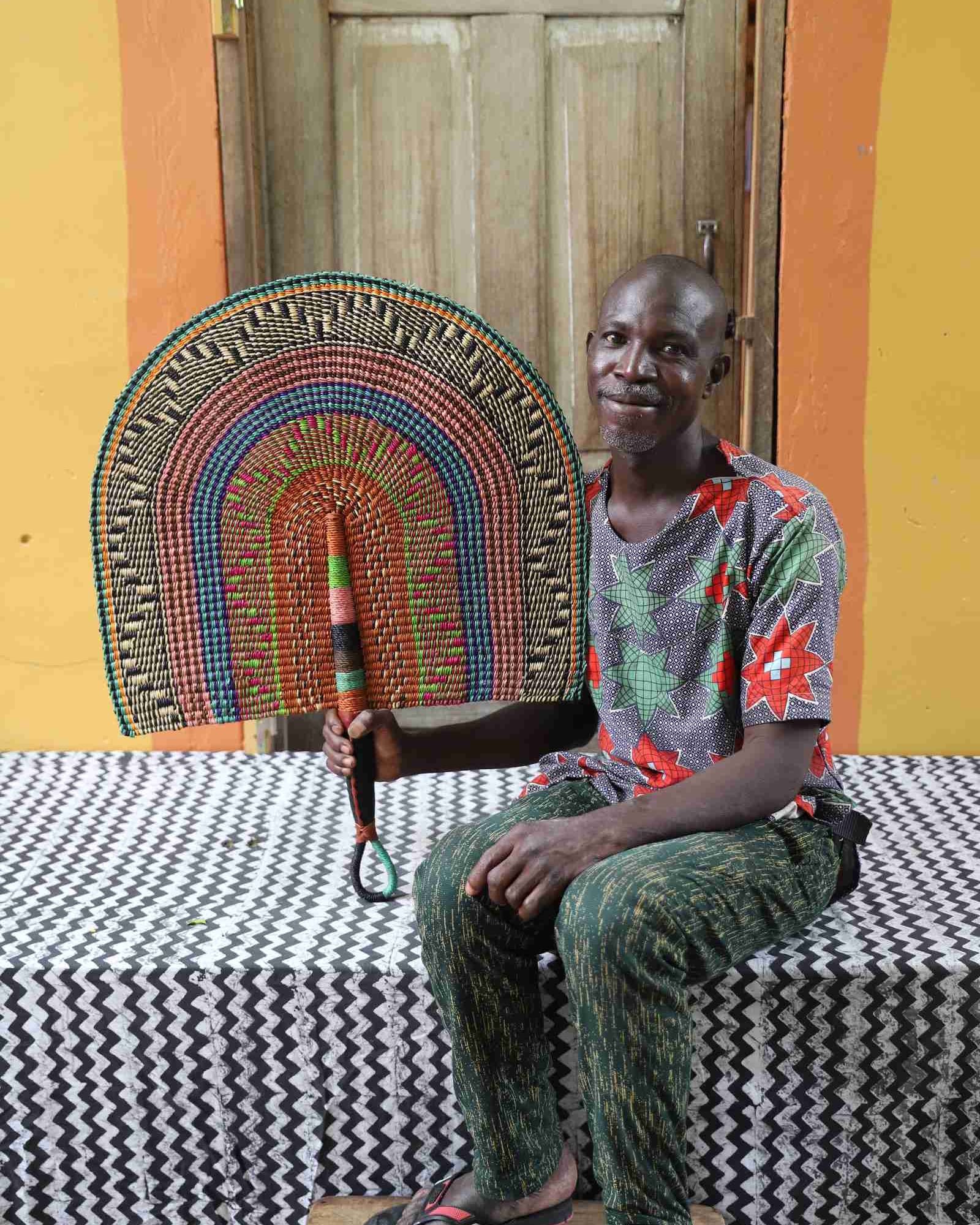 Naba Penga wall fan in natural straw with teal, rust and green multicolour bands, held by a seated person for scale.