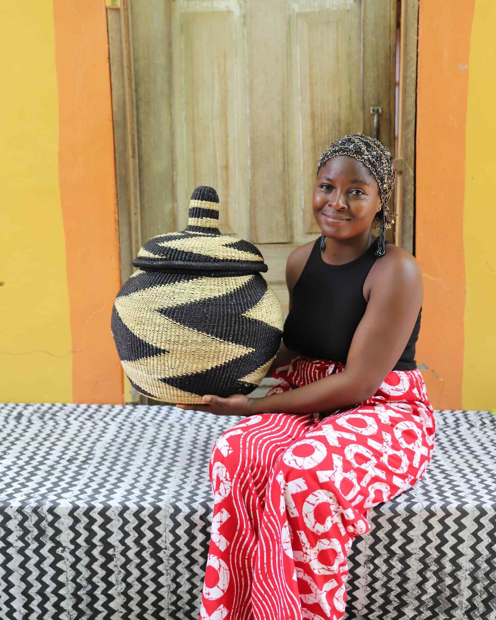 Lidded Bolga storage basket in a black and natural zigzag pattern, held by a seated woman for scale.