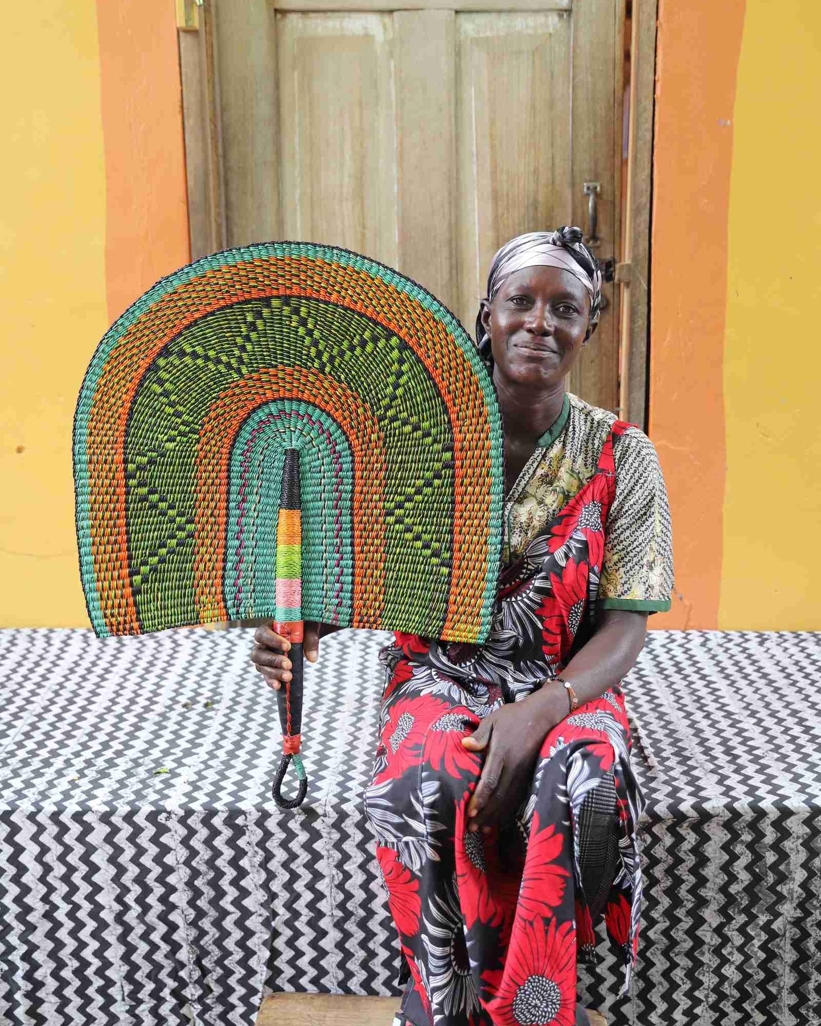 Naba Penga wall fan in green woven straw with an orange border, held by a seated person for scale.