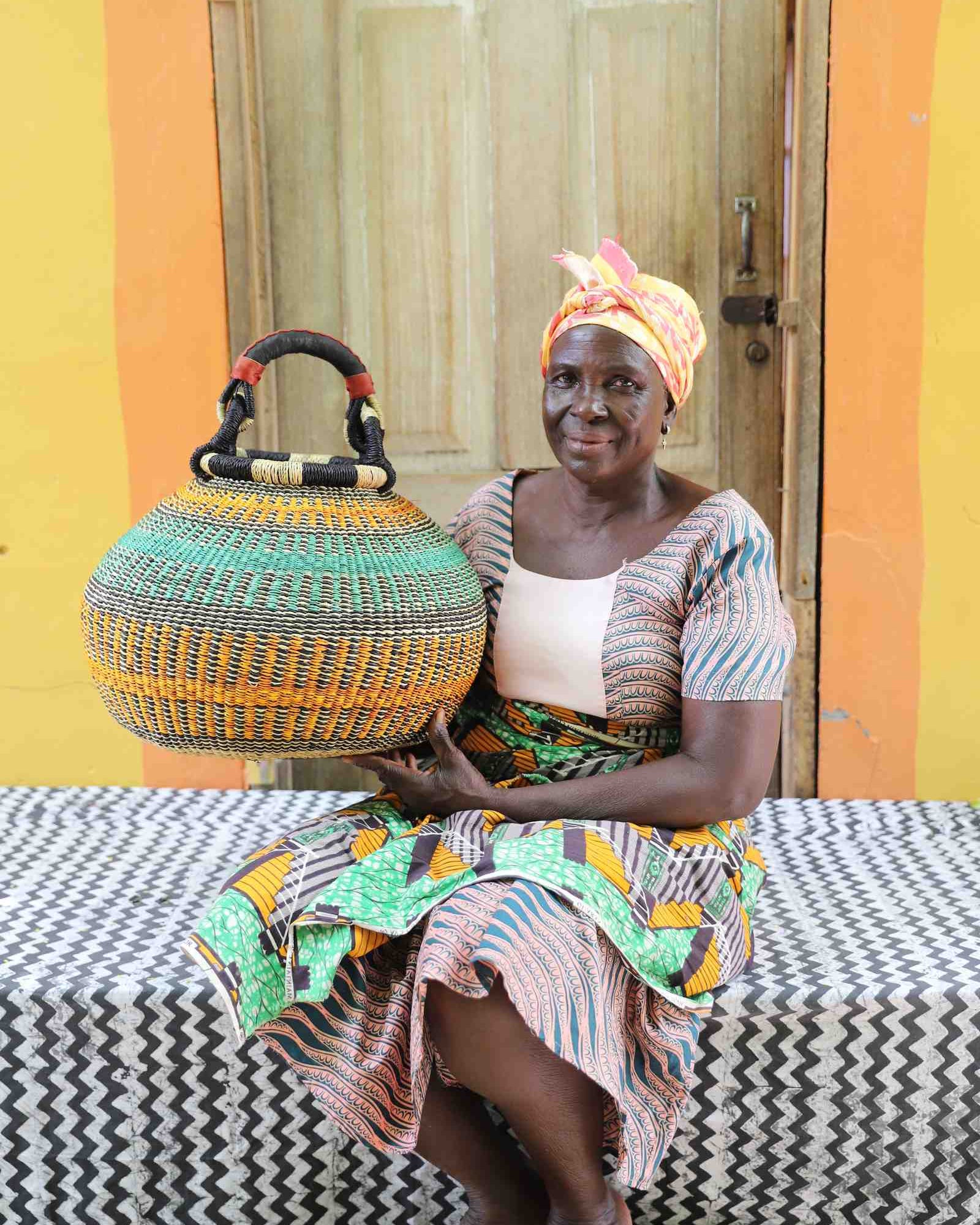 Round Bolga pot basket in orange and green stripes with a leather-wrapped handle, held by a seated woman.