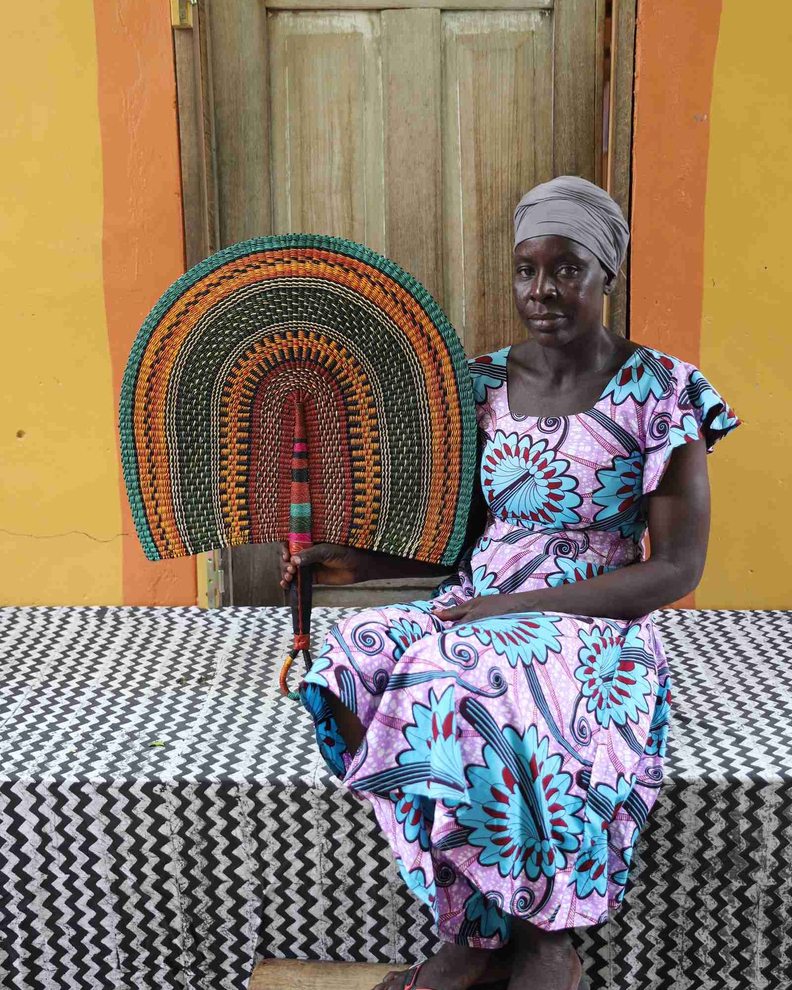 Naba Penga wall fan in natural straw with teal, orange and green concentric bands, held by a seated person for scale.