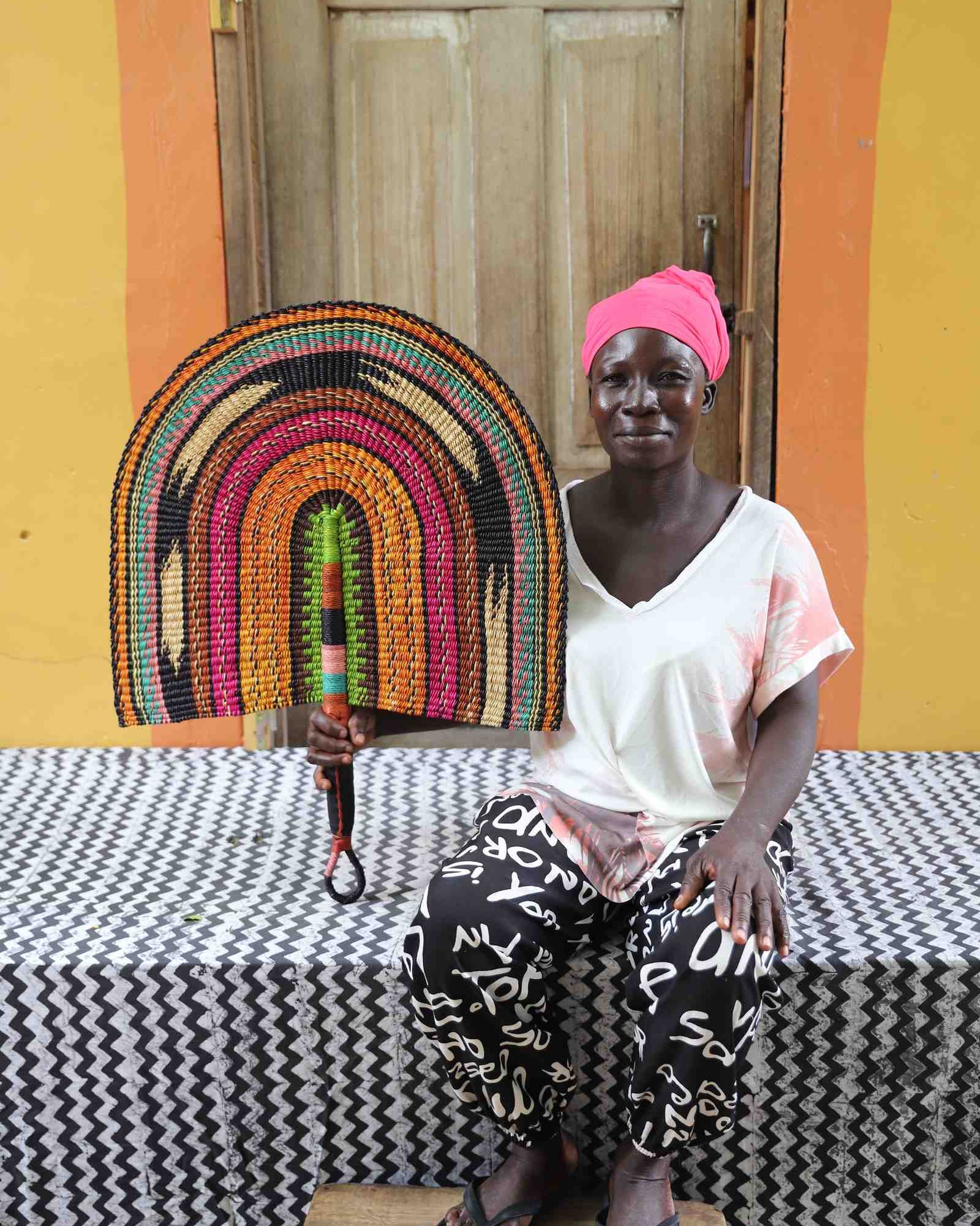 Naba Penga wall fan in natural straw with rust, green, pink and black rainbow arches, held by a seated person for scale.