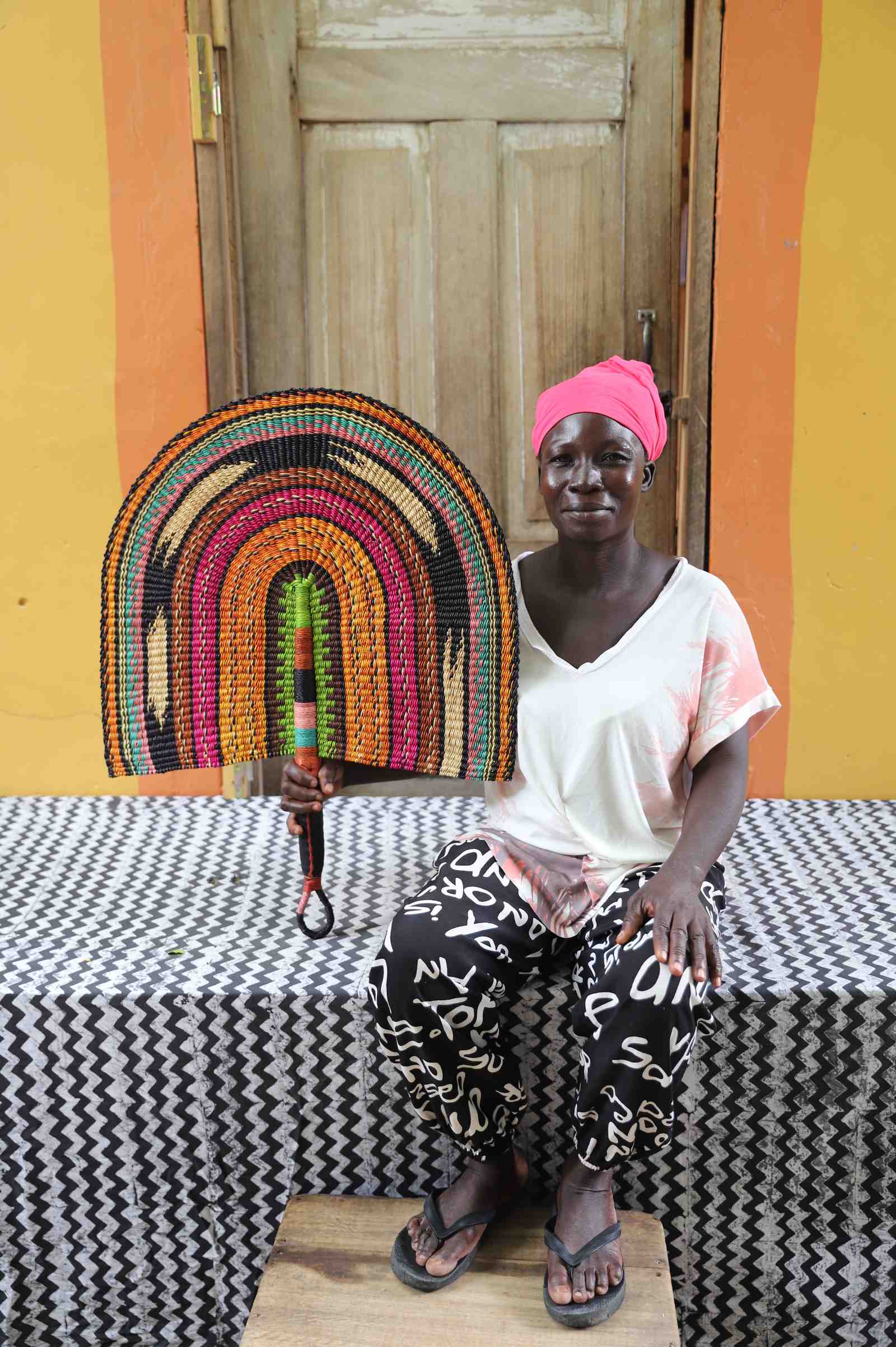 Naba Penga wall fan in natural straw with rust, green, pink and black rainbow arches, held by a seated person for scale.