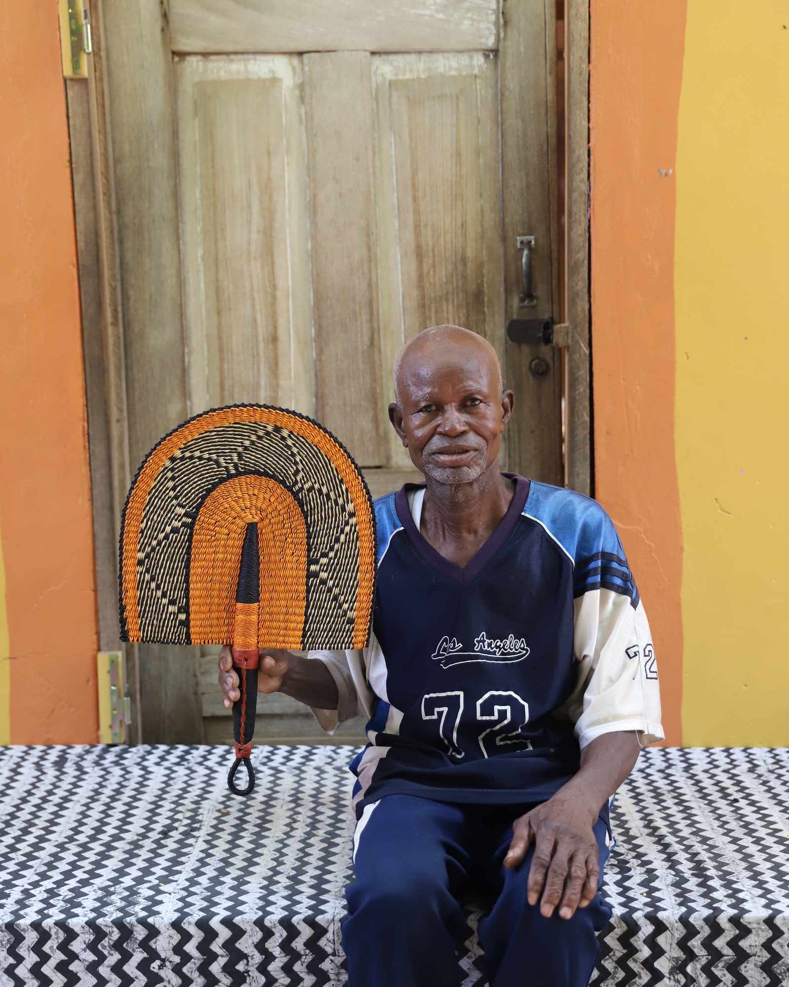 Weaver holding a handwoven Bolga Fan in natural straw with black geometric outer arch, in Bolgatanga, Ghana.