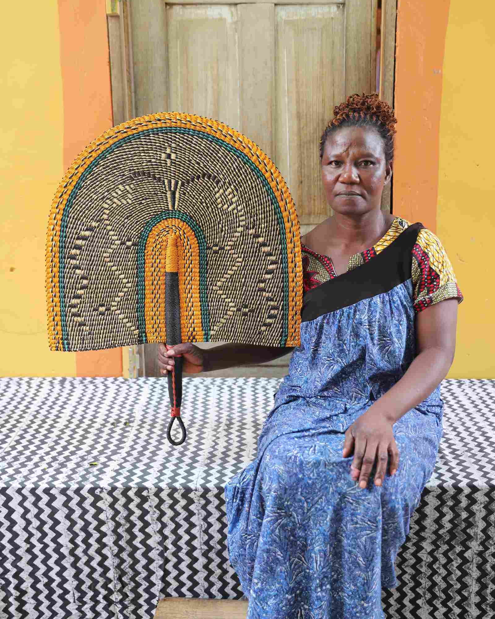 Naba Penga wall fan in natural straw with a mustard border and green trim, held by a seated person for scale.