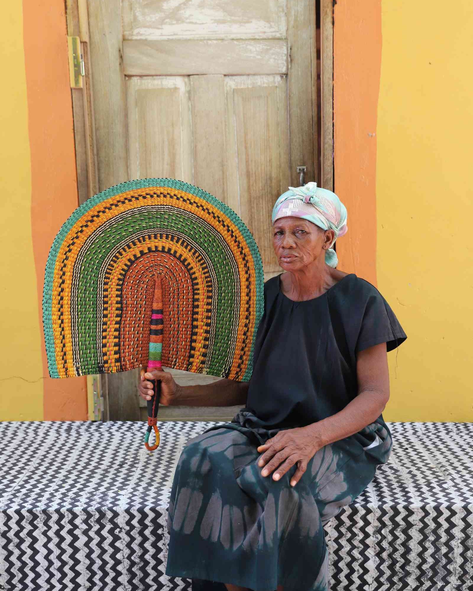 Weaver holding a Naba Penga handwoven wall fan with arch bands in green, amber and terracotta, in Bolgatanga, Ghana.