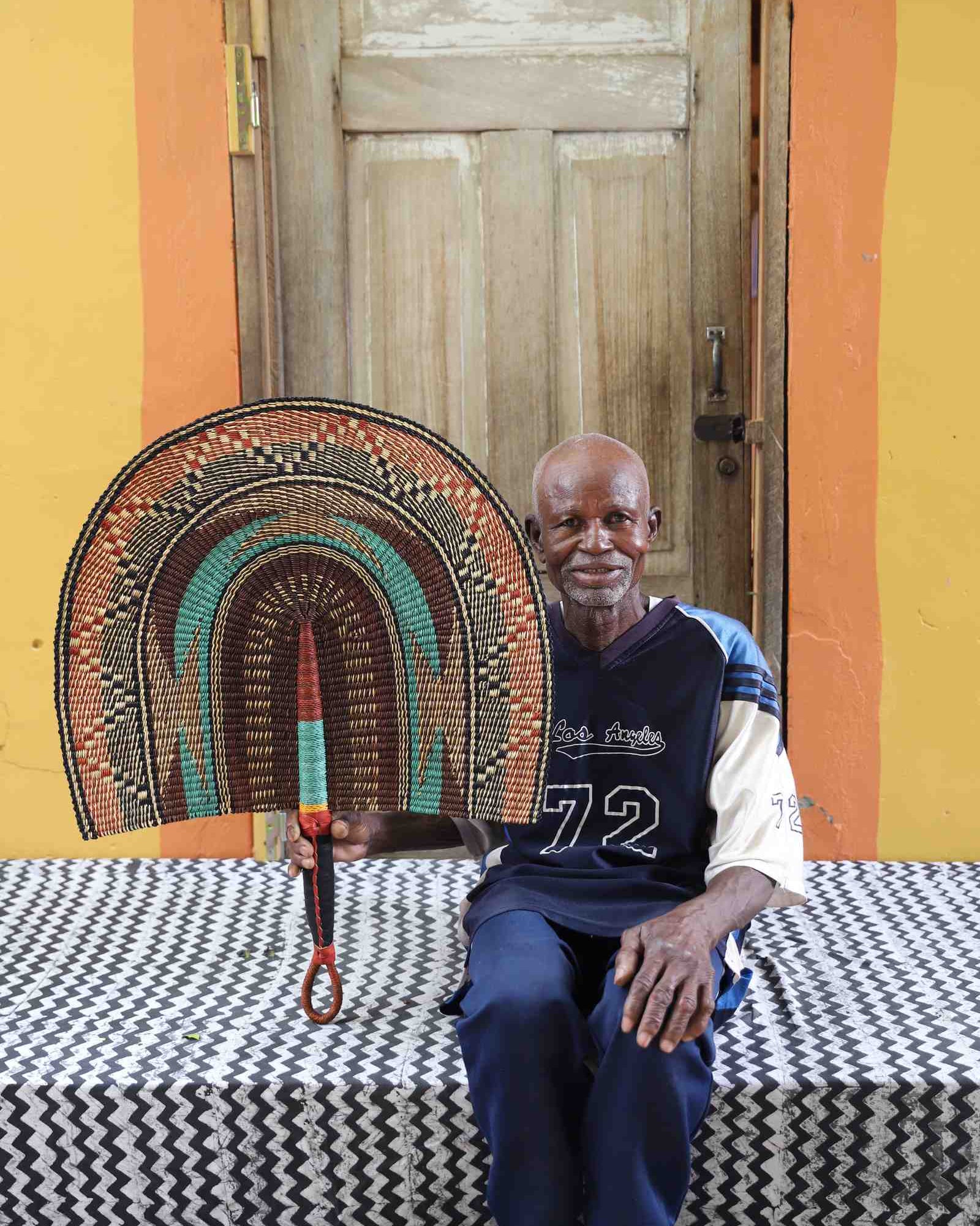 Naba Penga wall fan in natural straw with a teal inner arch and dark patterned border, held by a seated person for scale.