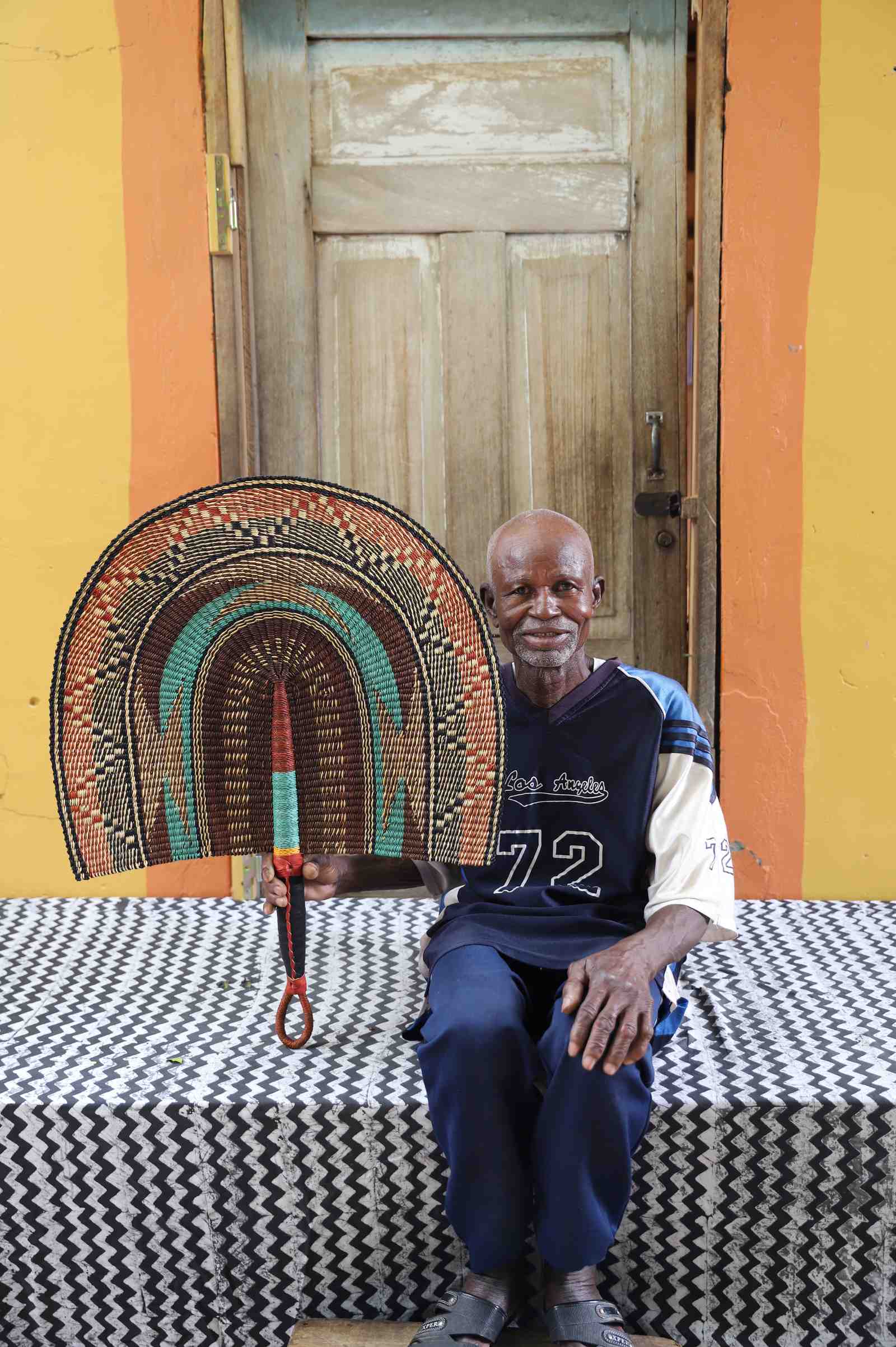 Naba Penga wall fan in natural straw with a teal inner arch and dark patterned border, held by a seated person for scale.