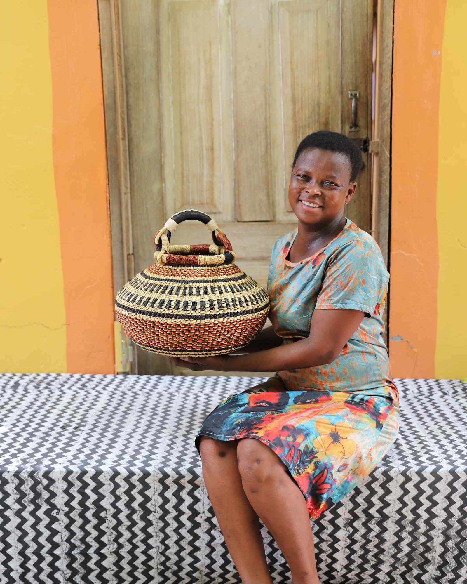 Small round Bolga pot basket in black and cream diamond weave with a rust band, held by a seated woman for scale.
