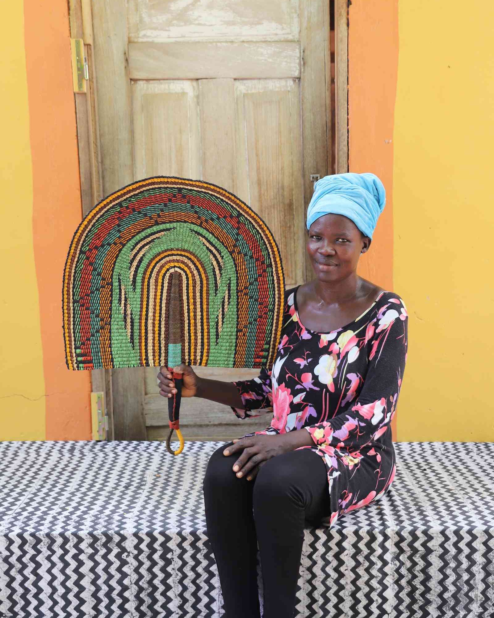 Weaver holding a Naba Penga handwoven wall fan with arch bands in green, terracotta and amber, in Bolgatanga, Ghana.